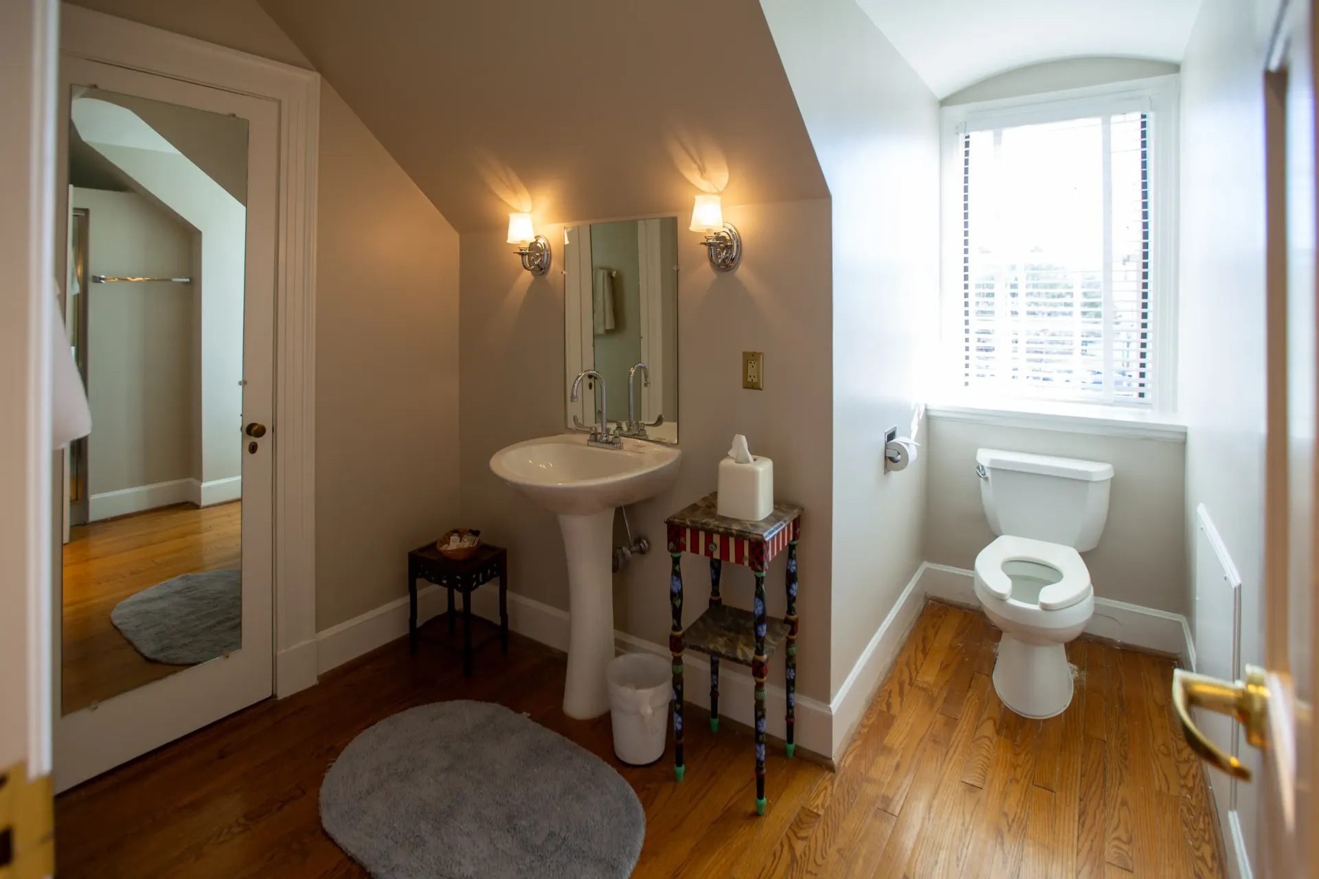 Bathroom with pedestal sink, toilet, window, and wooden floor.