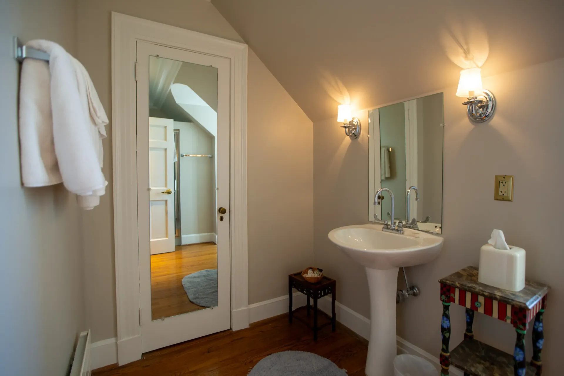 Bathroom with pedestal sink, mirror, and a small side table. Light-colored walls and wood floor.
