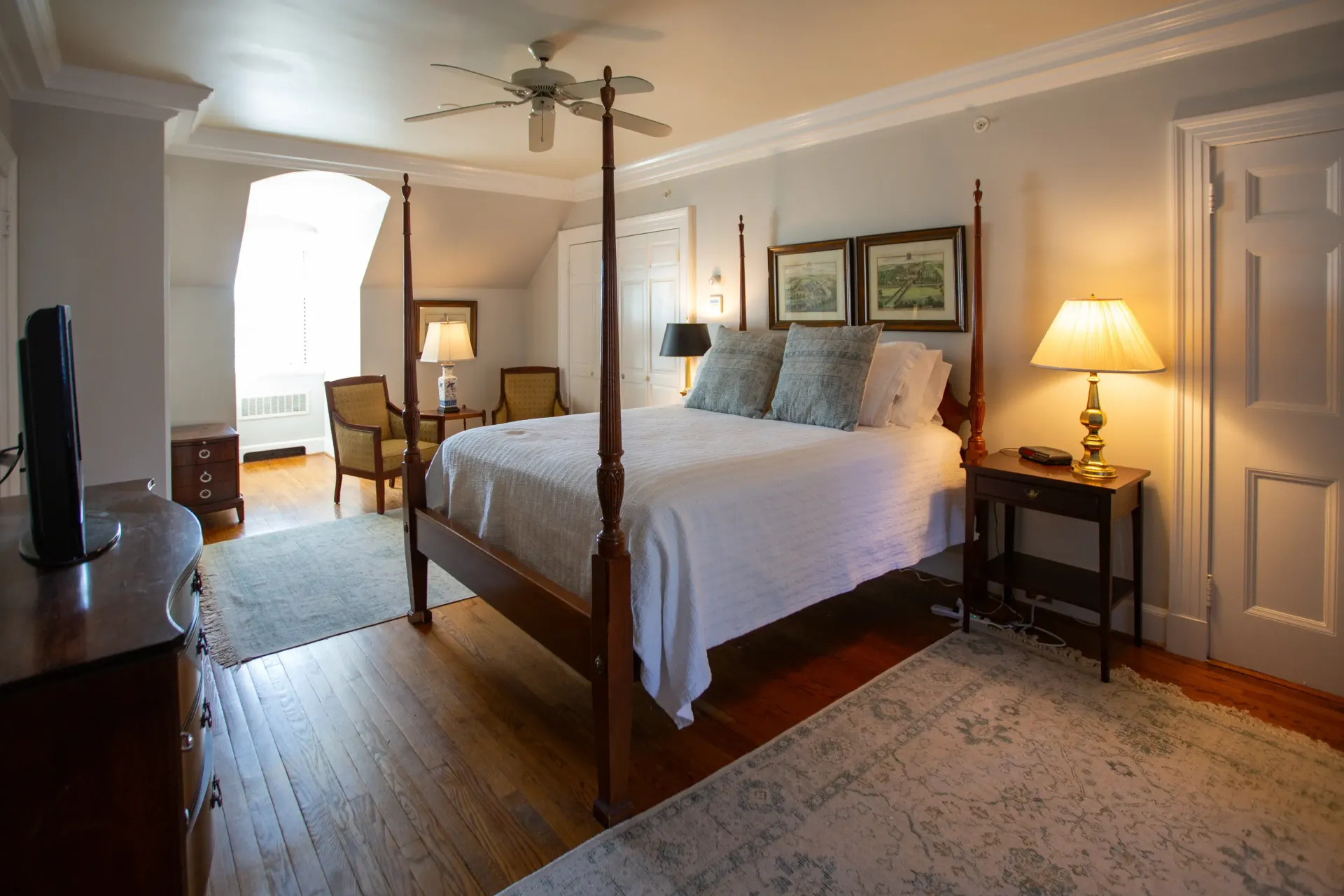 Bedroom with a four-poster bed, rug, lamps, and a TV. The room is painted white.