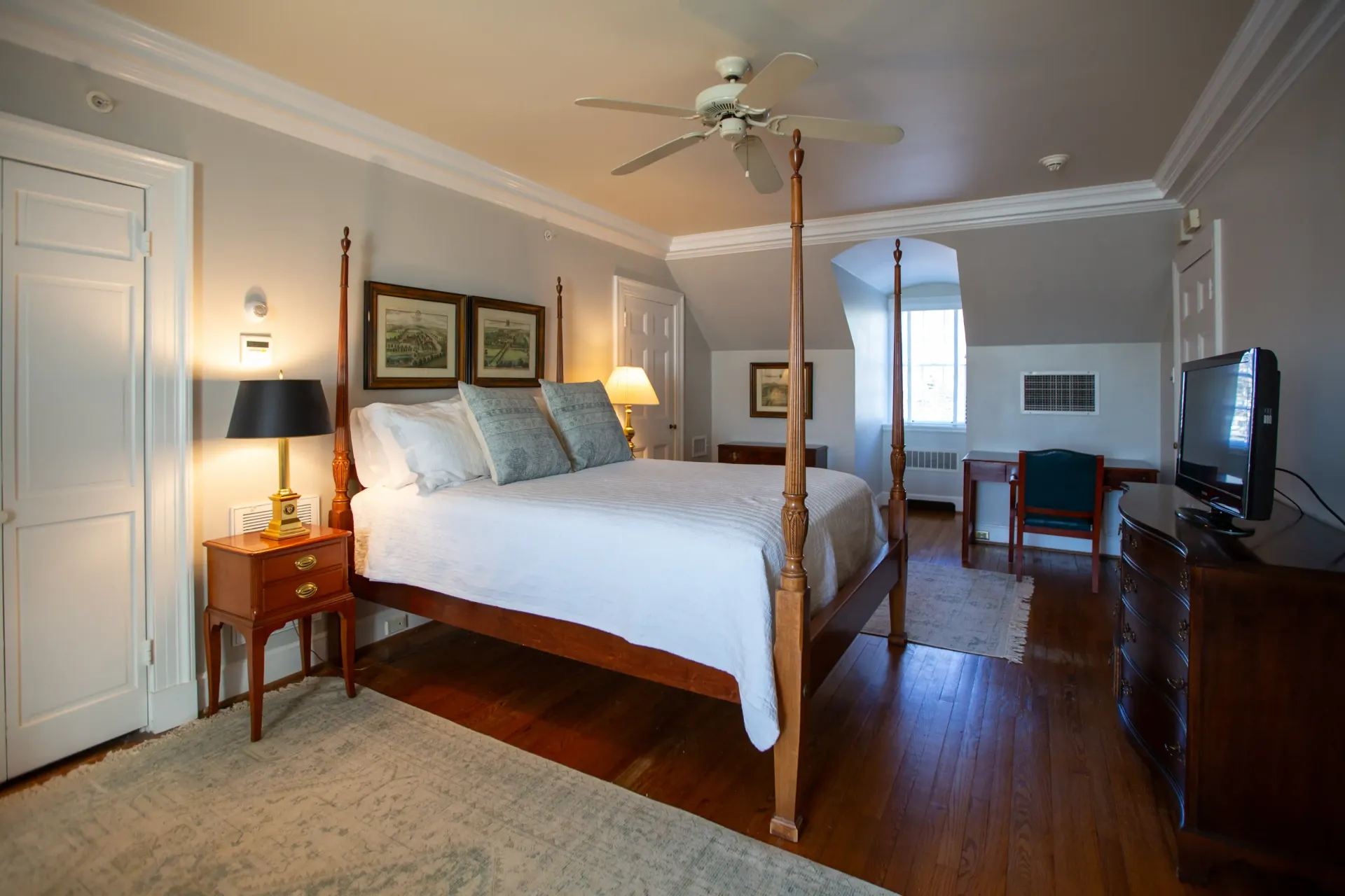 Bedroom with four-poster bed, nightstand, dresser, and desk; light gray walls, wood floor, and ceiling fan.