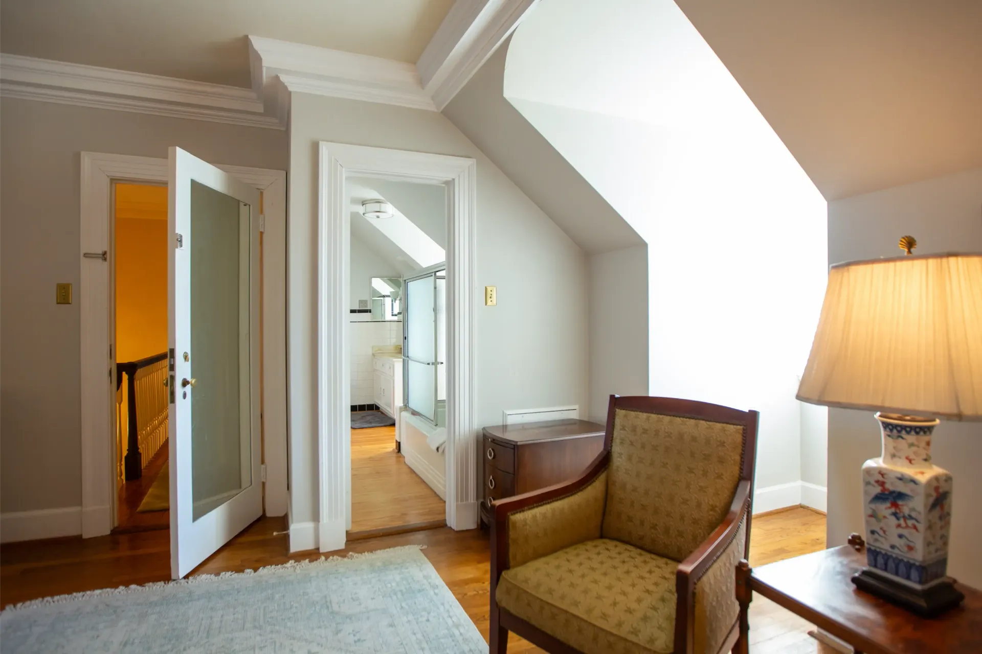 Cozy attic room with a chair, lamp, and doors leading to a staircase and a bathroom.
