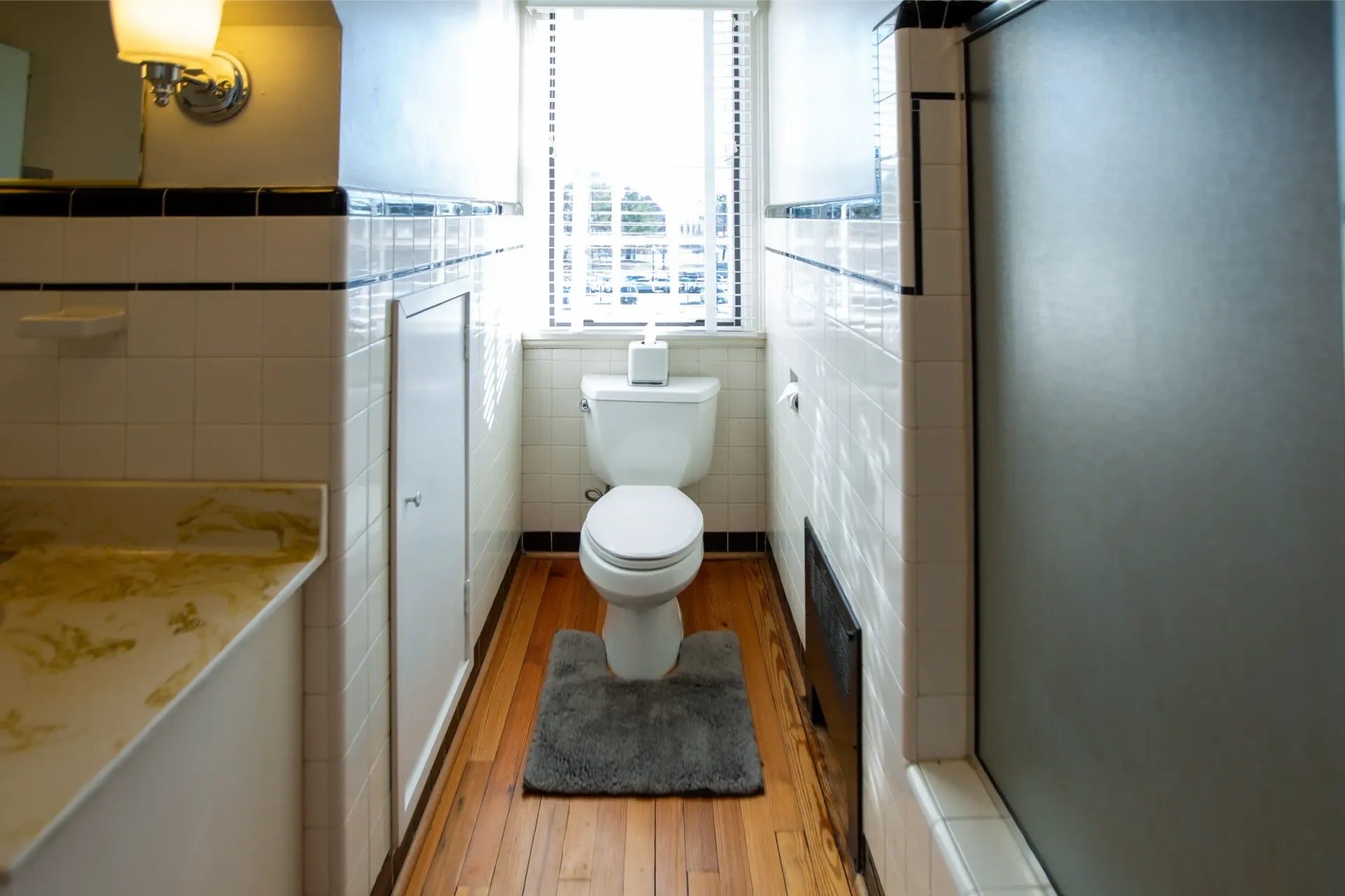 Narrow bathroom with white tile walls, toilet, and wooden floor. Window with blinds and grey bath mat.