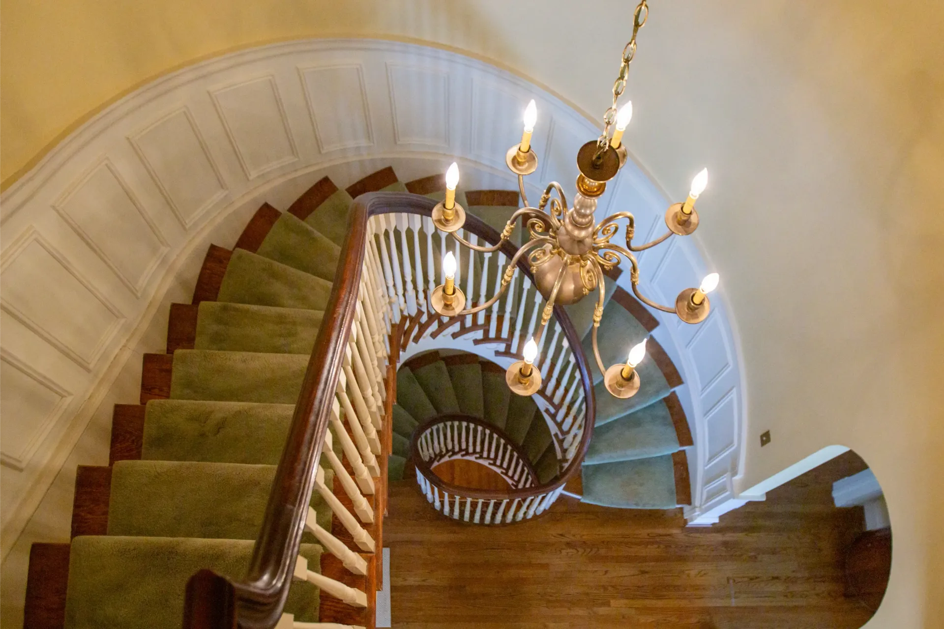 Spiral staircase with green carpet, wooden railing, and chandelier.