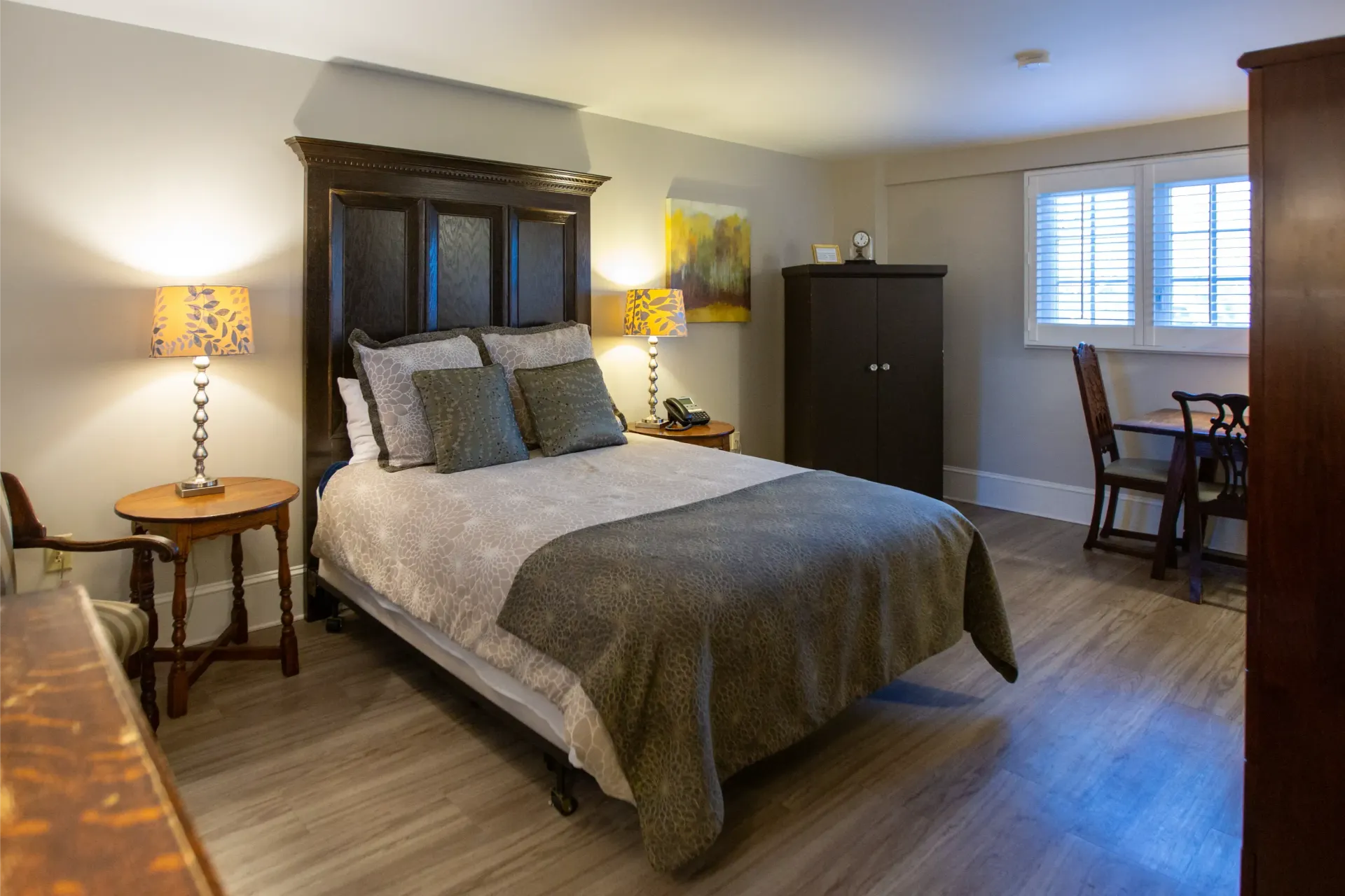 Bedroom with a dark wooden bed frame, bedside tables, a wardrobe, and a small table and chairs near a window.