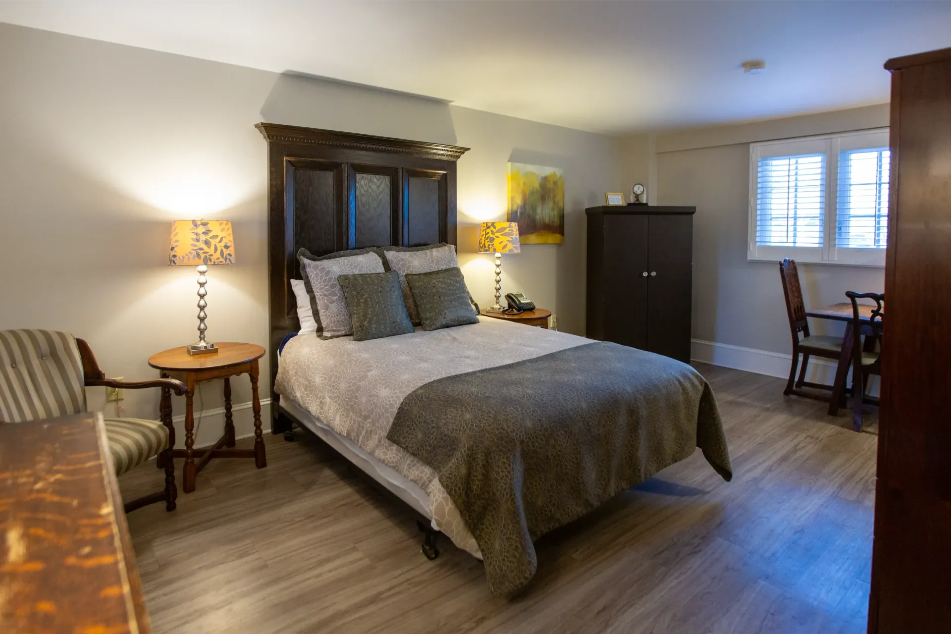 Bedroom with bed, nightstands, wardrobe, and small dining table. Soft lighting with wood flooring and neutral walls.
