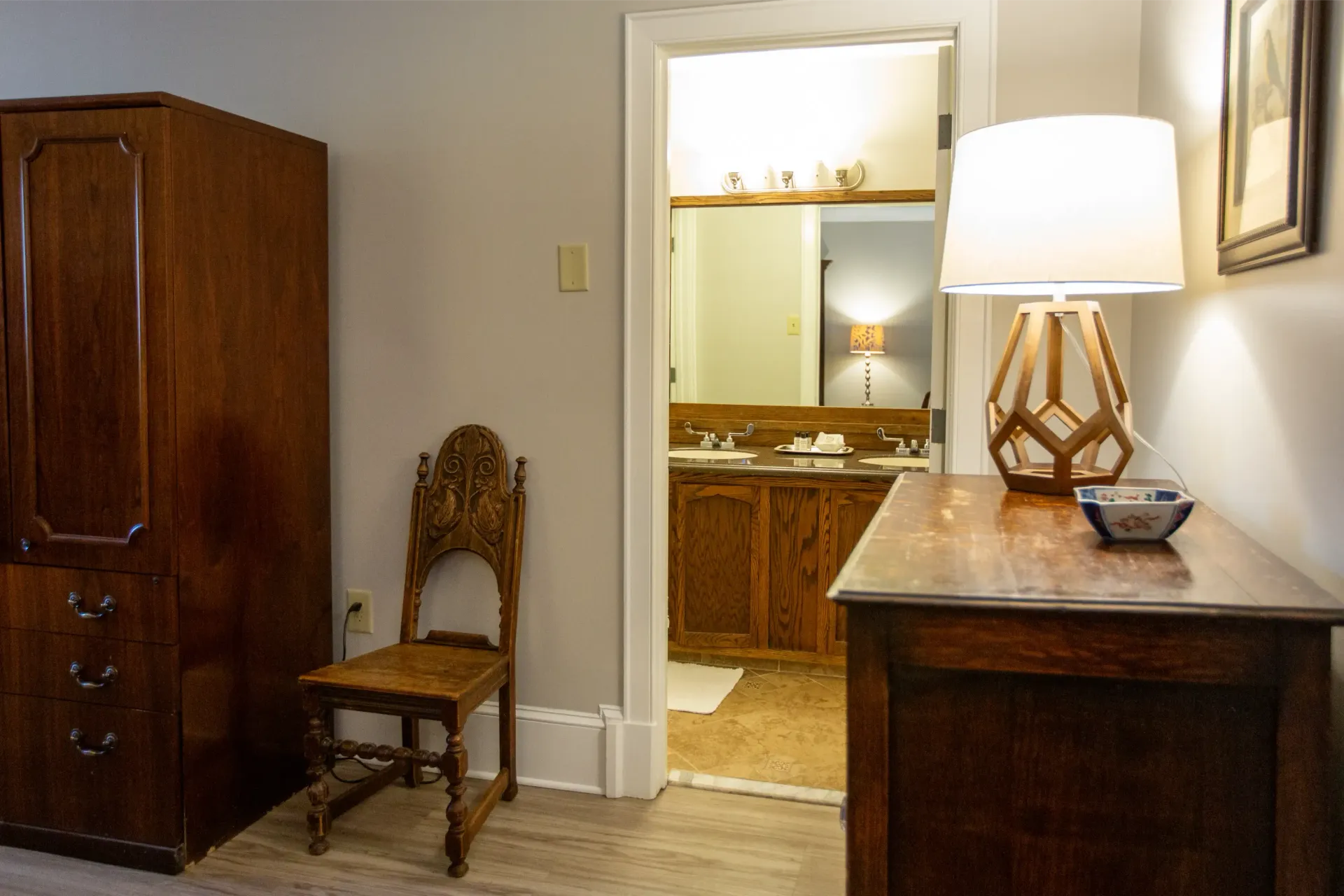 Bedroom interior with wooden furniture, including a wardrobe, chair, and dresser. Bathroom visible through doorway.