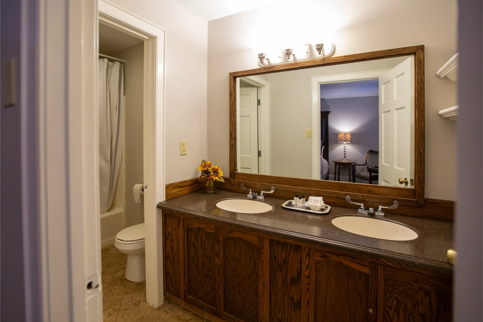 Bathroom with a large wooden vanity, double sinks, a mirror, and a view into the bedroom.