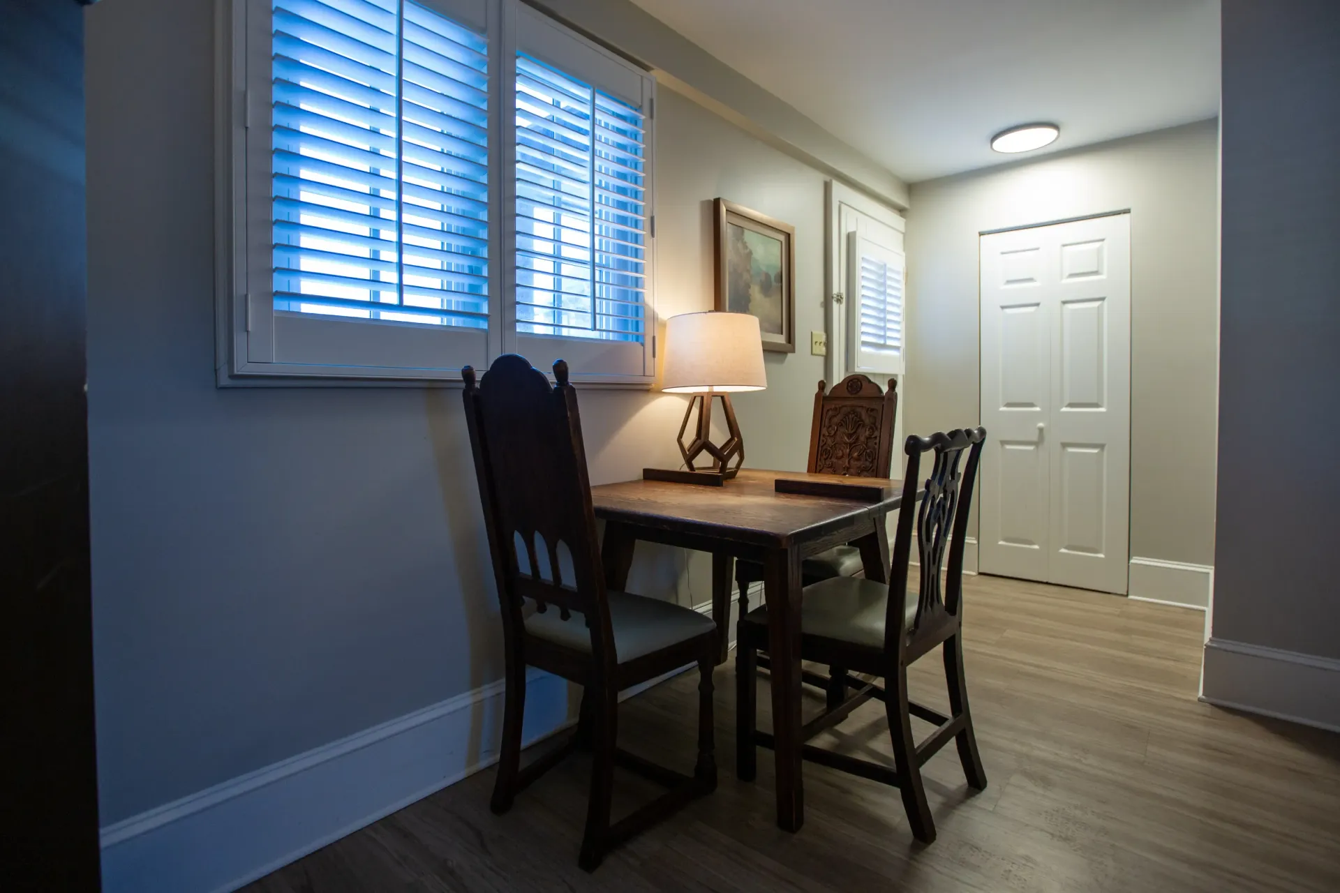 A small dining area with a table, chairs, lamp, and window with blinds. Light gray walls, light wooden floor.