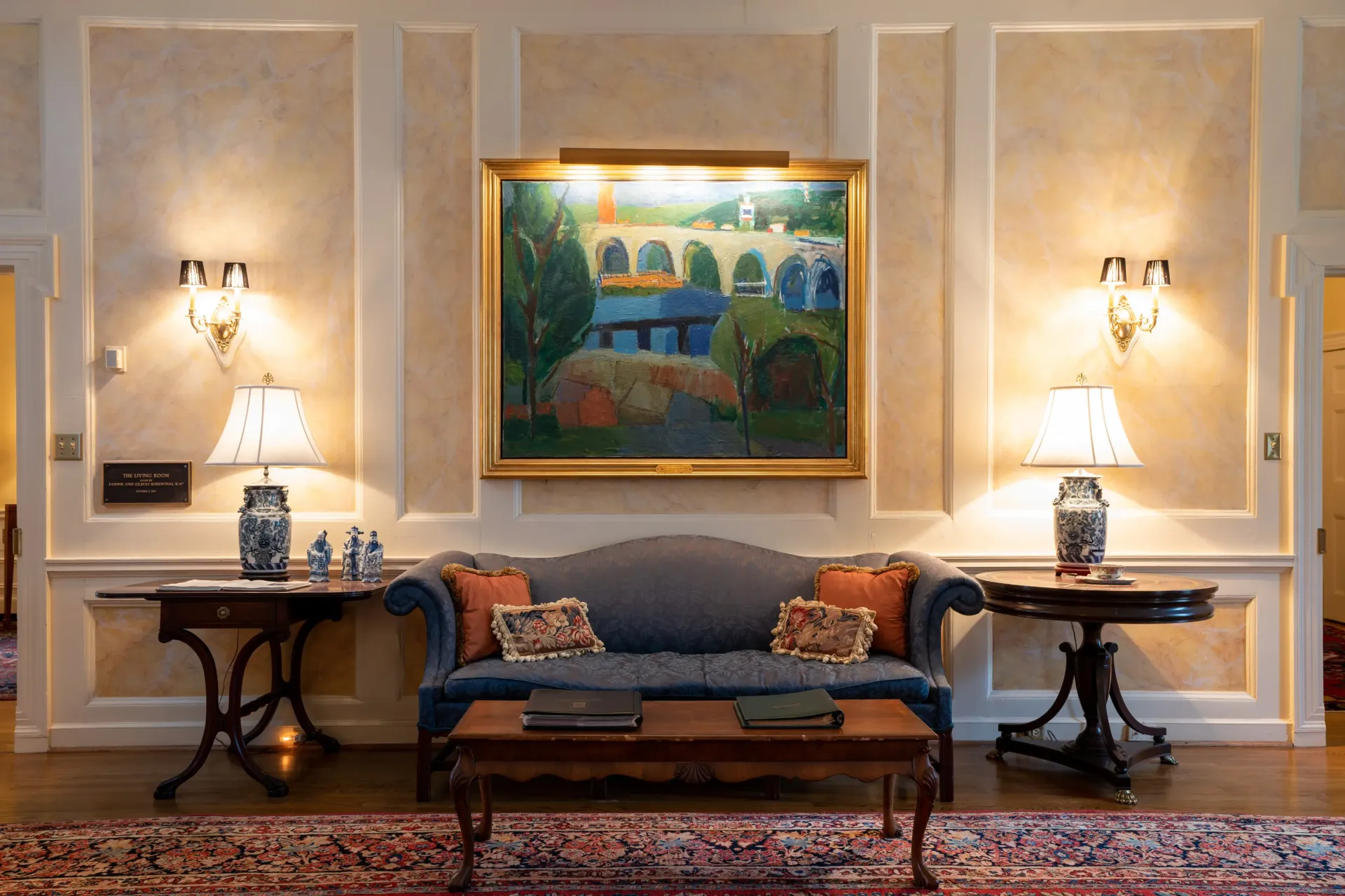 Elegant room with a blue sofa, artwork, lamps, and tables. Beige walls with white trim, warm lighting.