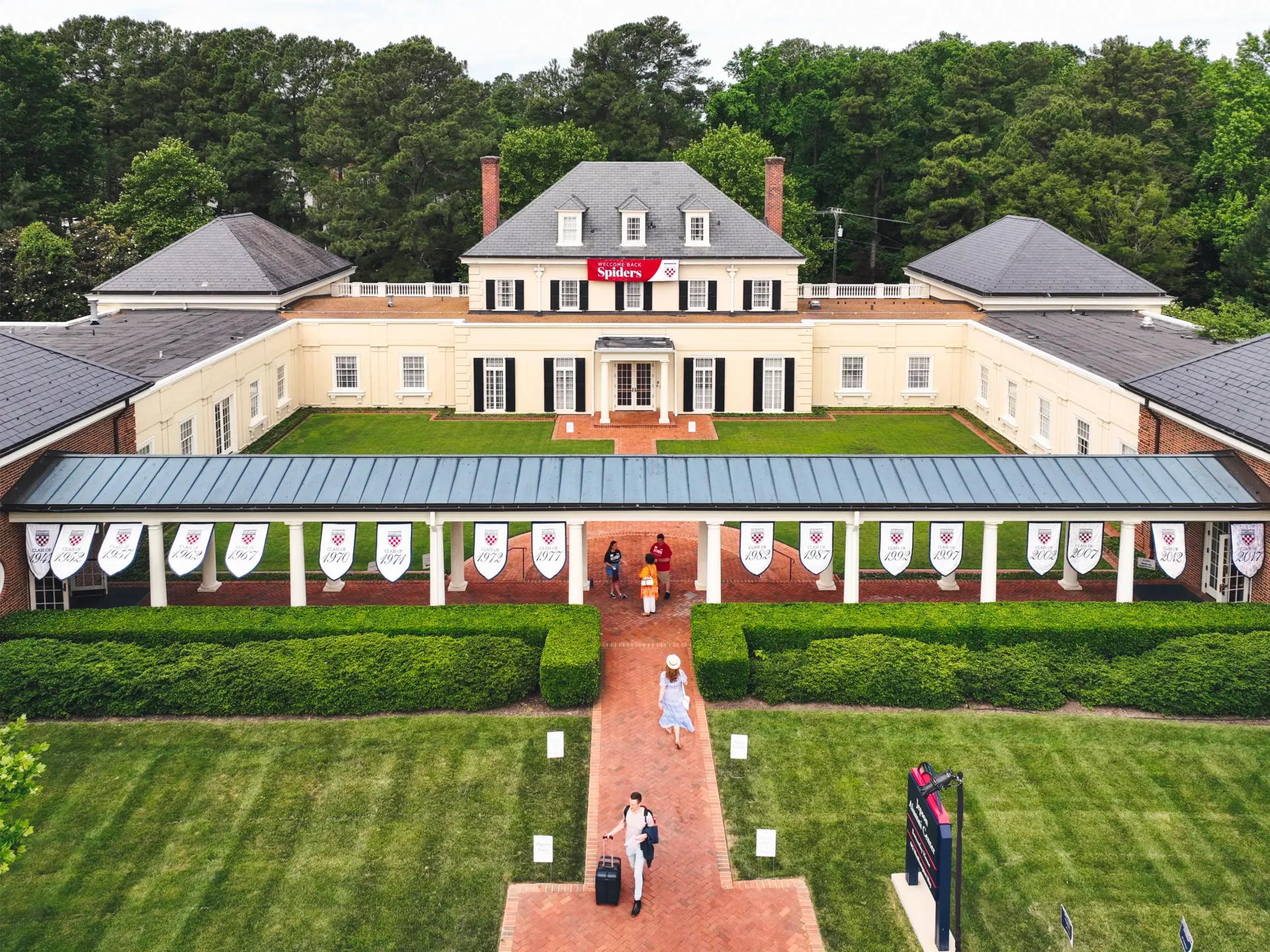 Exterior shot of a large, cream-colored building with manicured lawn and walkway; people walking toward the entrance.