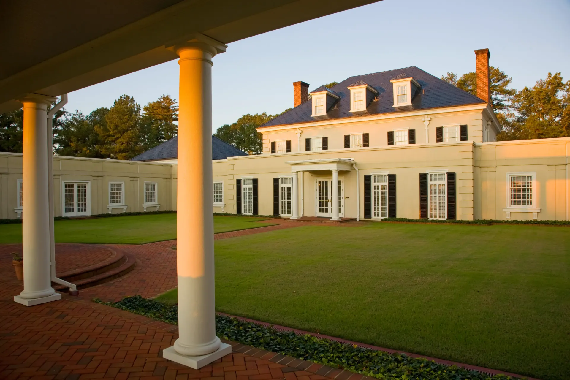 White columned porch of a light-colored mansion with black shutters and a manicured lawn.