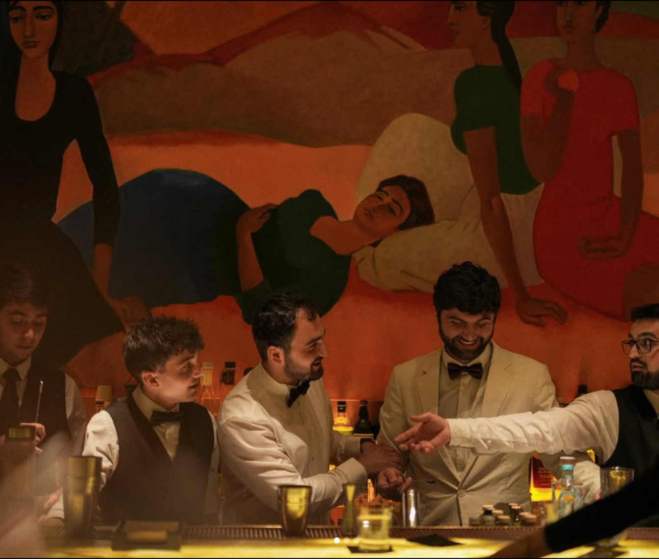 Bartenders behind a bar in front of a mural. One man shows his watch. Dark setting, warm colors.