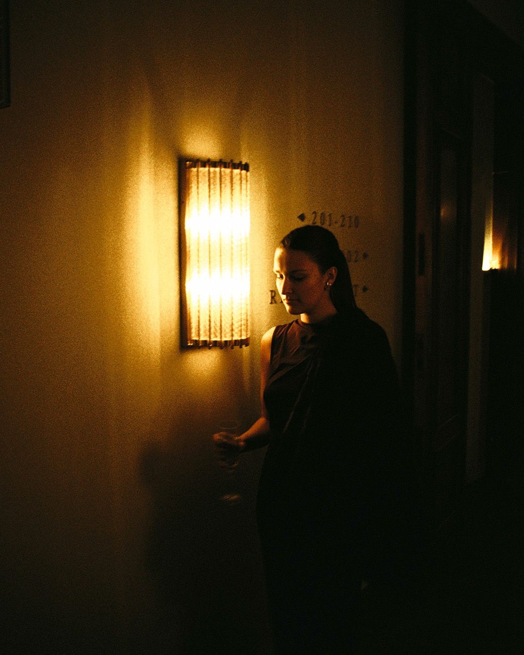 Bartenders behind a bar in front of a mural. One man shows his watch. Dark setting, warm colors.