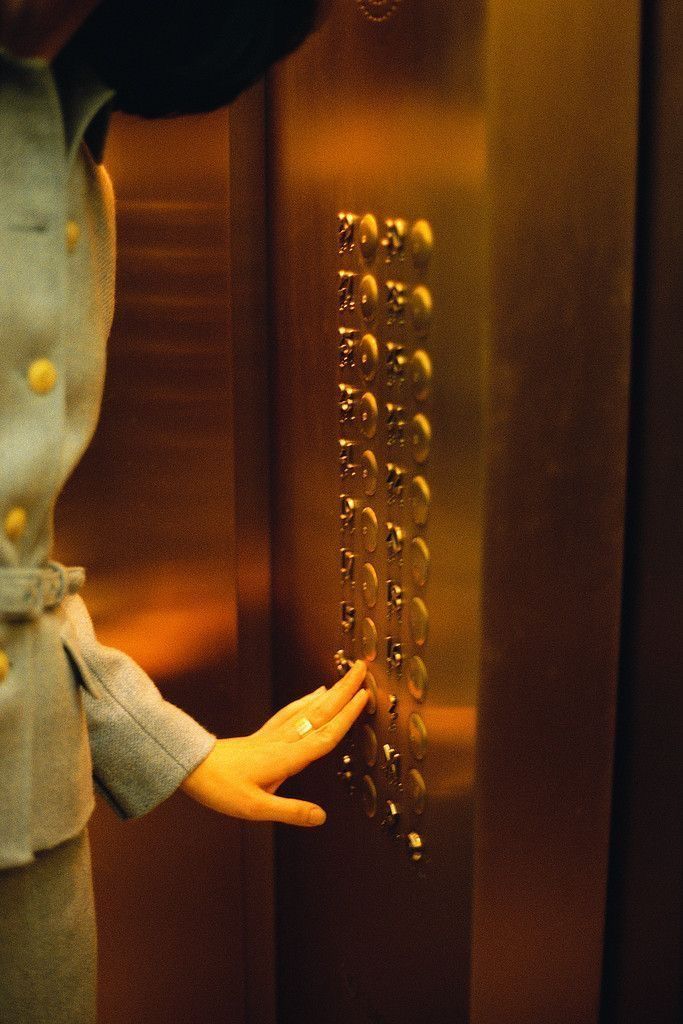 Woman's hand pressing elevator button on gold-colored panel, wearing a light blue suit.