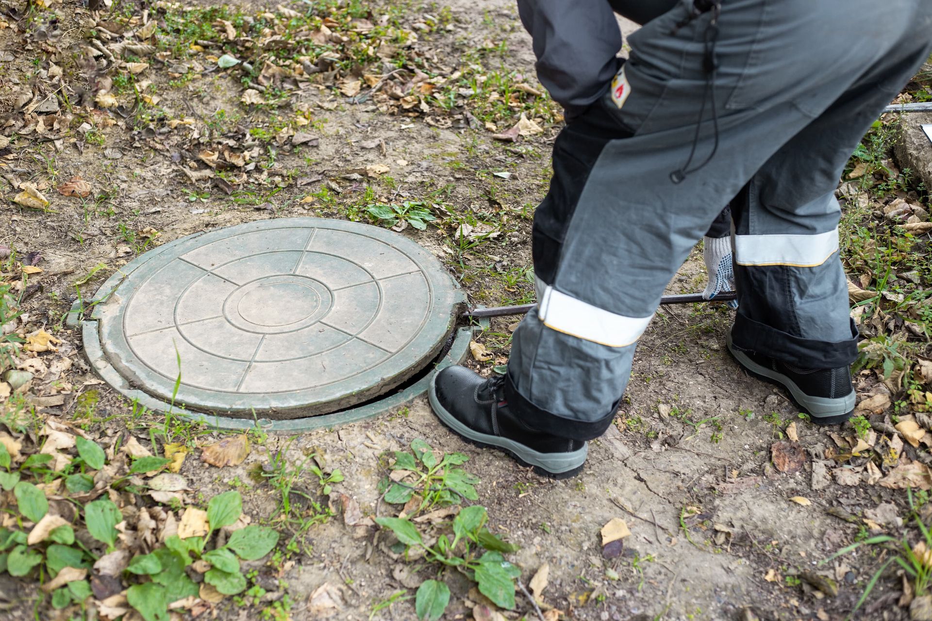 A person in work trousers and boots uses a metal tool to lift a circular concrete manhole cover from the ground.