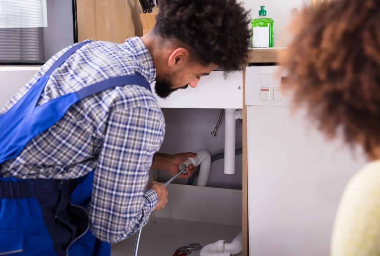 A plumber wearing a blue uniform works under a kitchen sink while a person looks on nearby.
