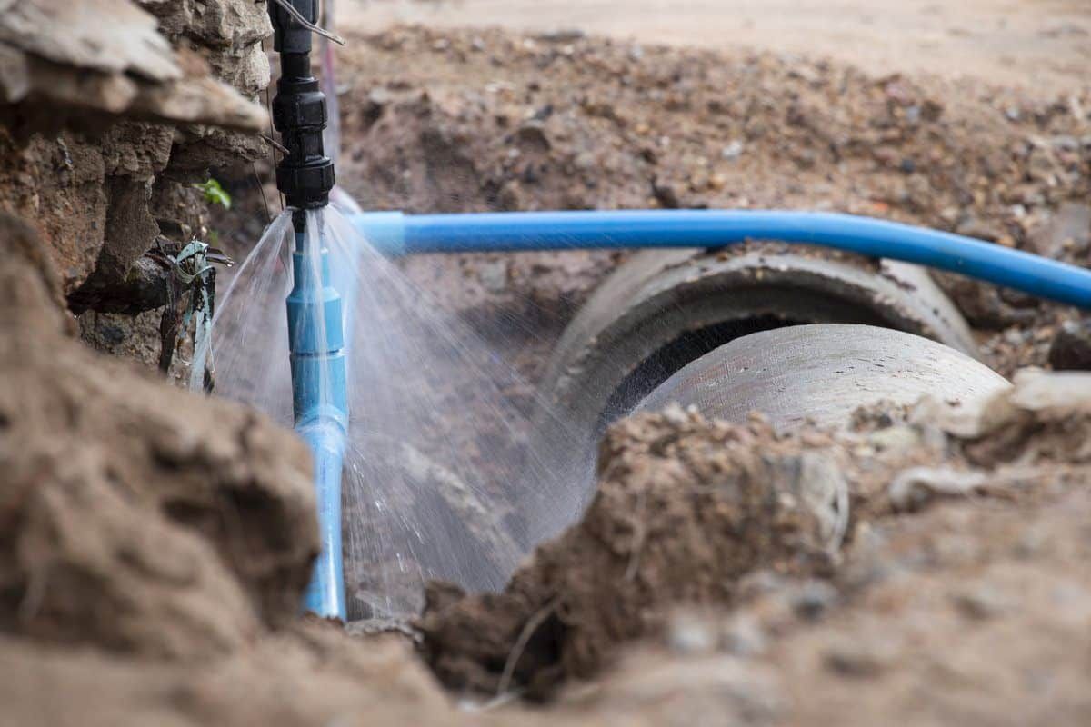 A blue plastic pipe buried in the ground is spraying water from a leaking joint.