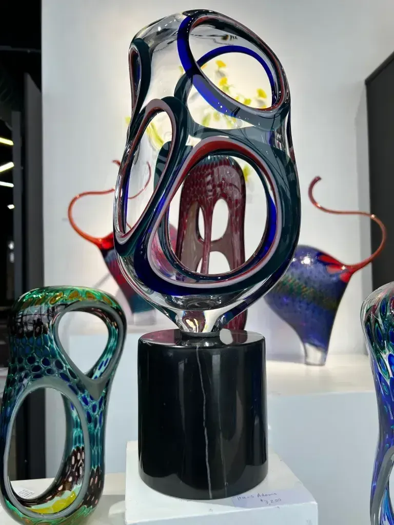 Sculptural glass art display; dark blue, red, and clear organic forms on black pedestals, in a gallery setting.