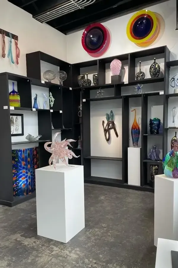 Art gallery with glass sculptures on pedestals and shelves; colorful glass art on walls.