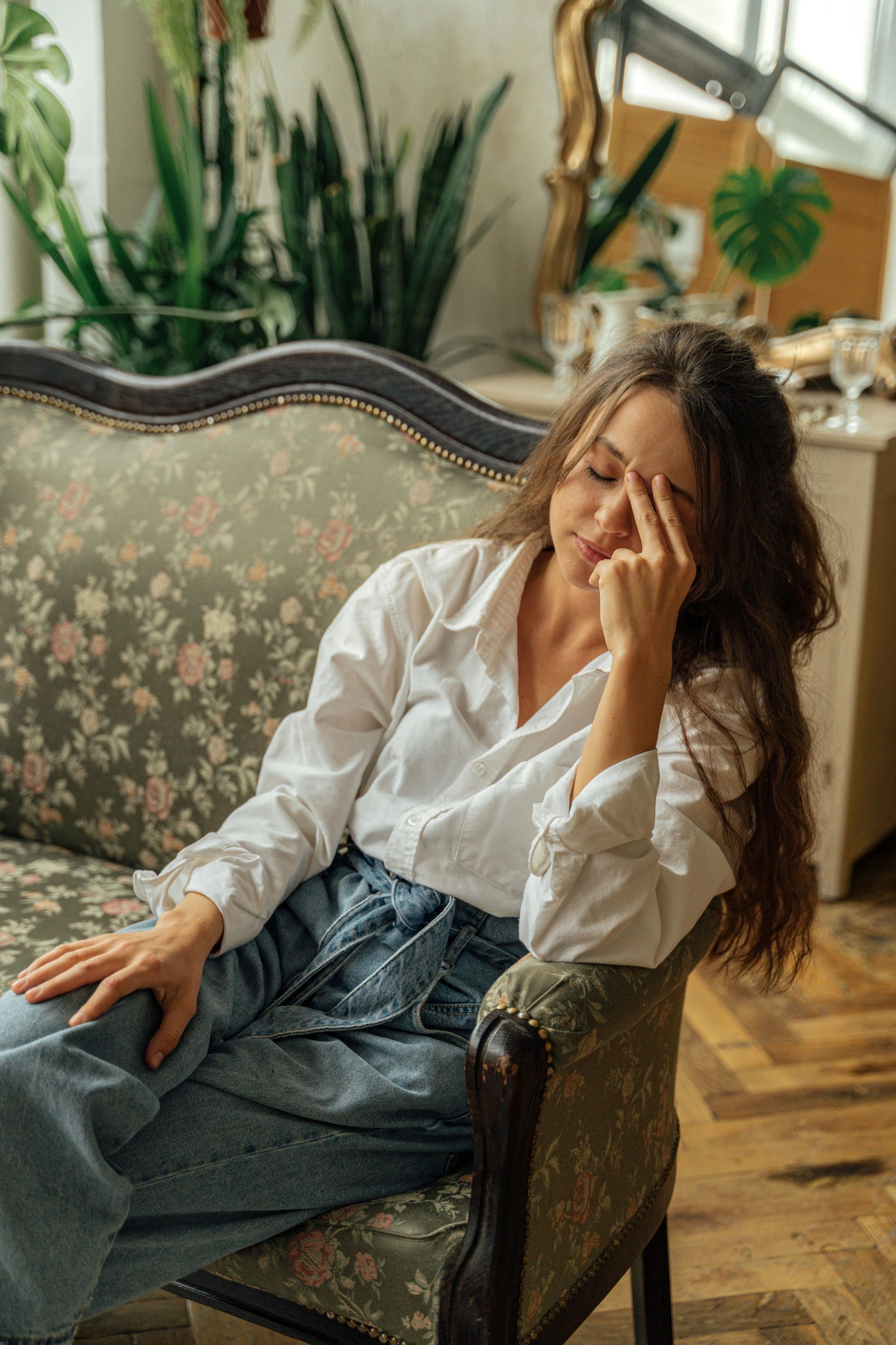 Woman in white shirt and jeans sits on floral couch, touching her forehead.