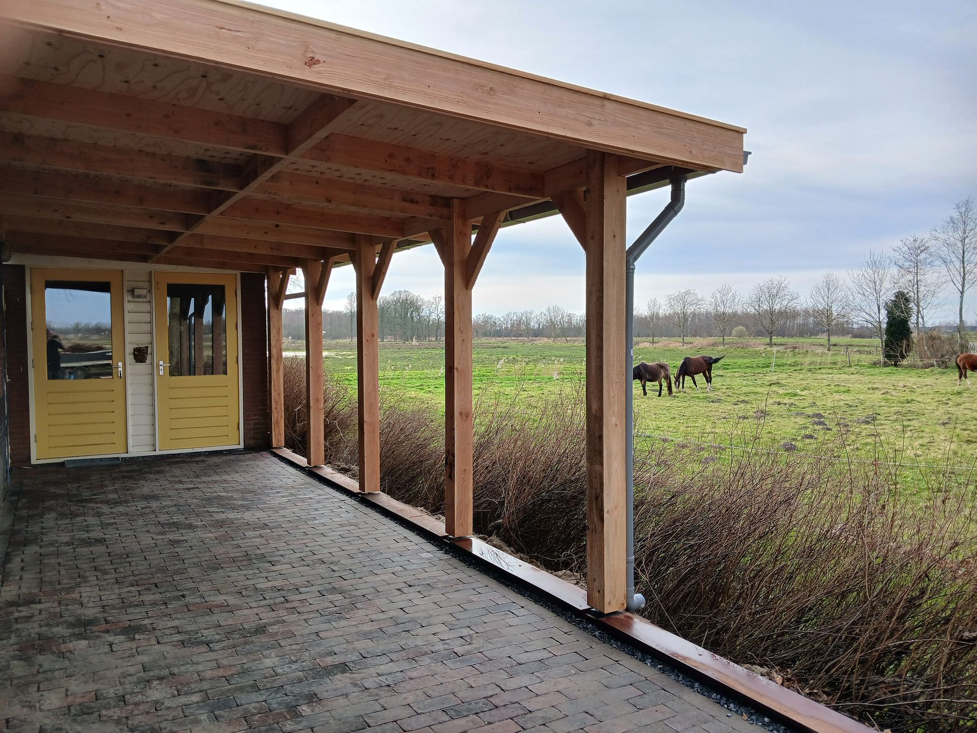 Overdekte veranda met uitzicht op een veld met grazende dieren. Houten palen en dak, deuren en groen.