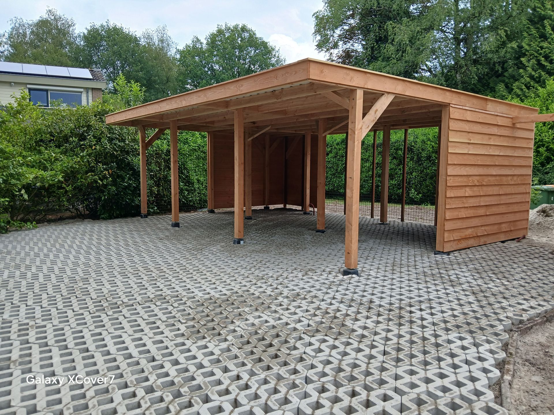 Houten carport op een grijs, rastervormig oppervlak, omgeven door groen.
