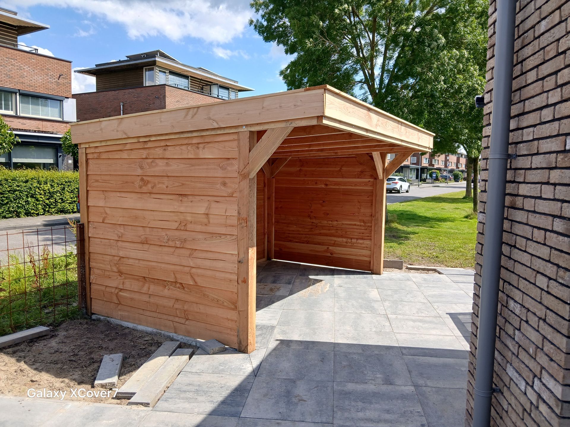 Houten carport met een plat dak, gelegen op een bestraat terrein naast een bakstenen gebouw en een straat.