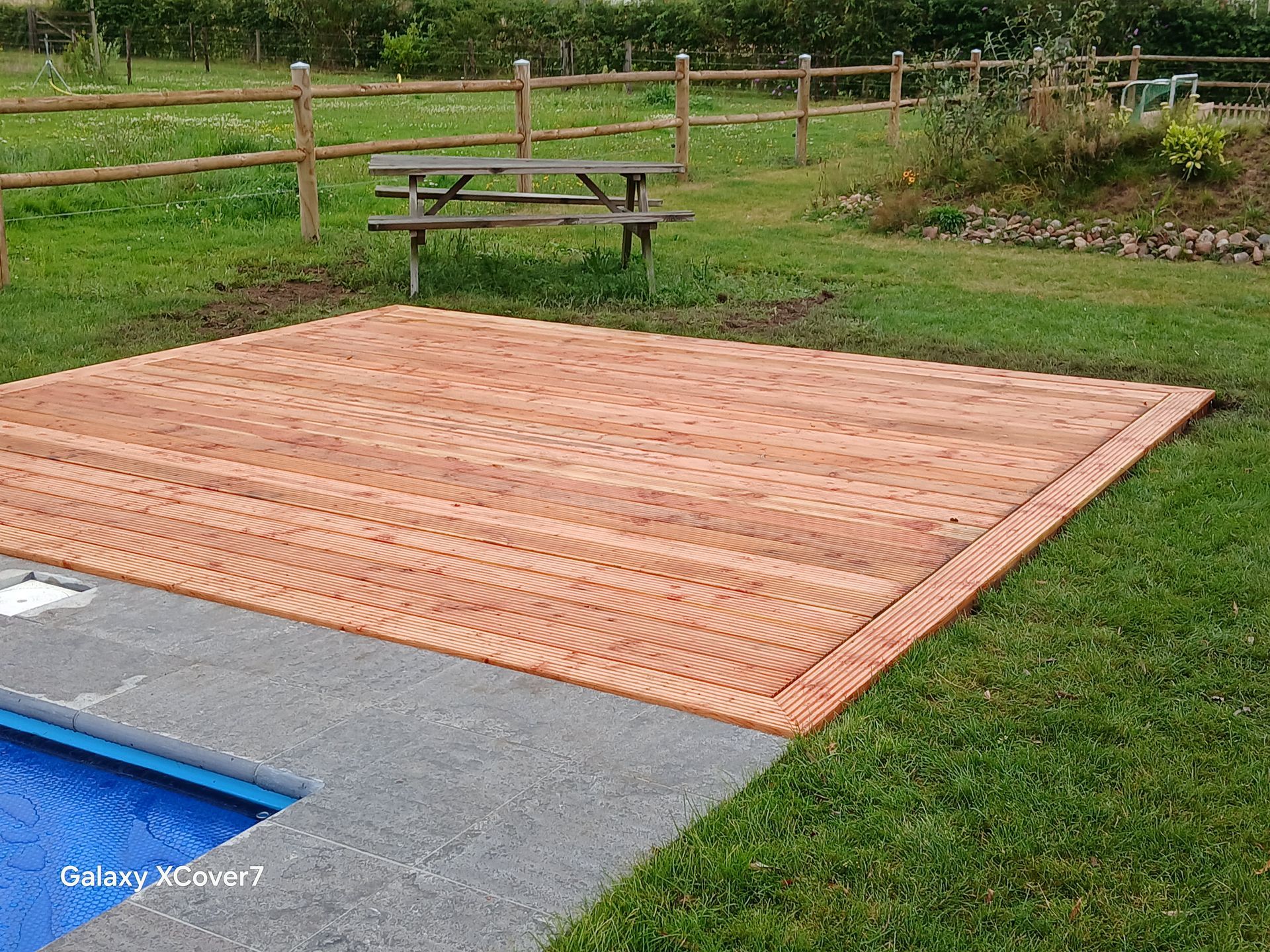 Zwembad met een bruin houten terras, gazon, picknicktafel en hek op de achtergrond.