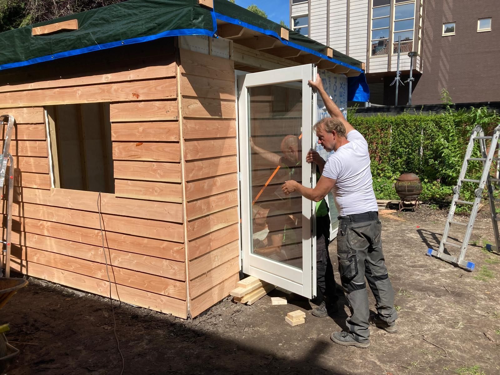 Een man installeert een witte deur in een houten schuur in een achtertuin.