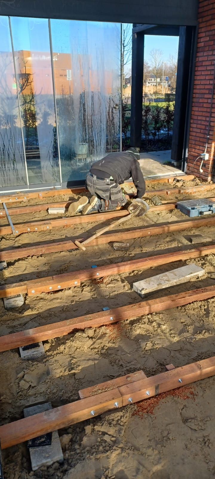 Een persoon knielt bij houten balken en glazen deuren tijdens de aanleg van een buitenterras.