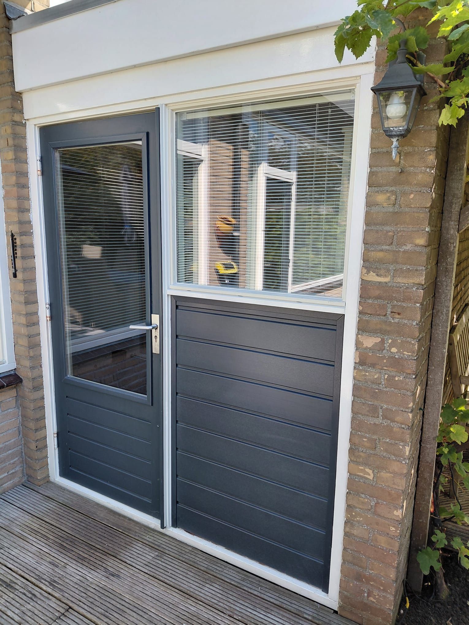 Donkergrijze deur en raam met horizontale jaloezieën, bakstenen muur en buitenverlichting.