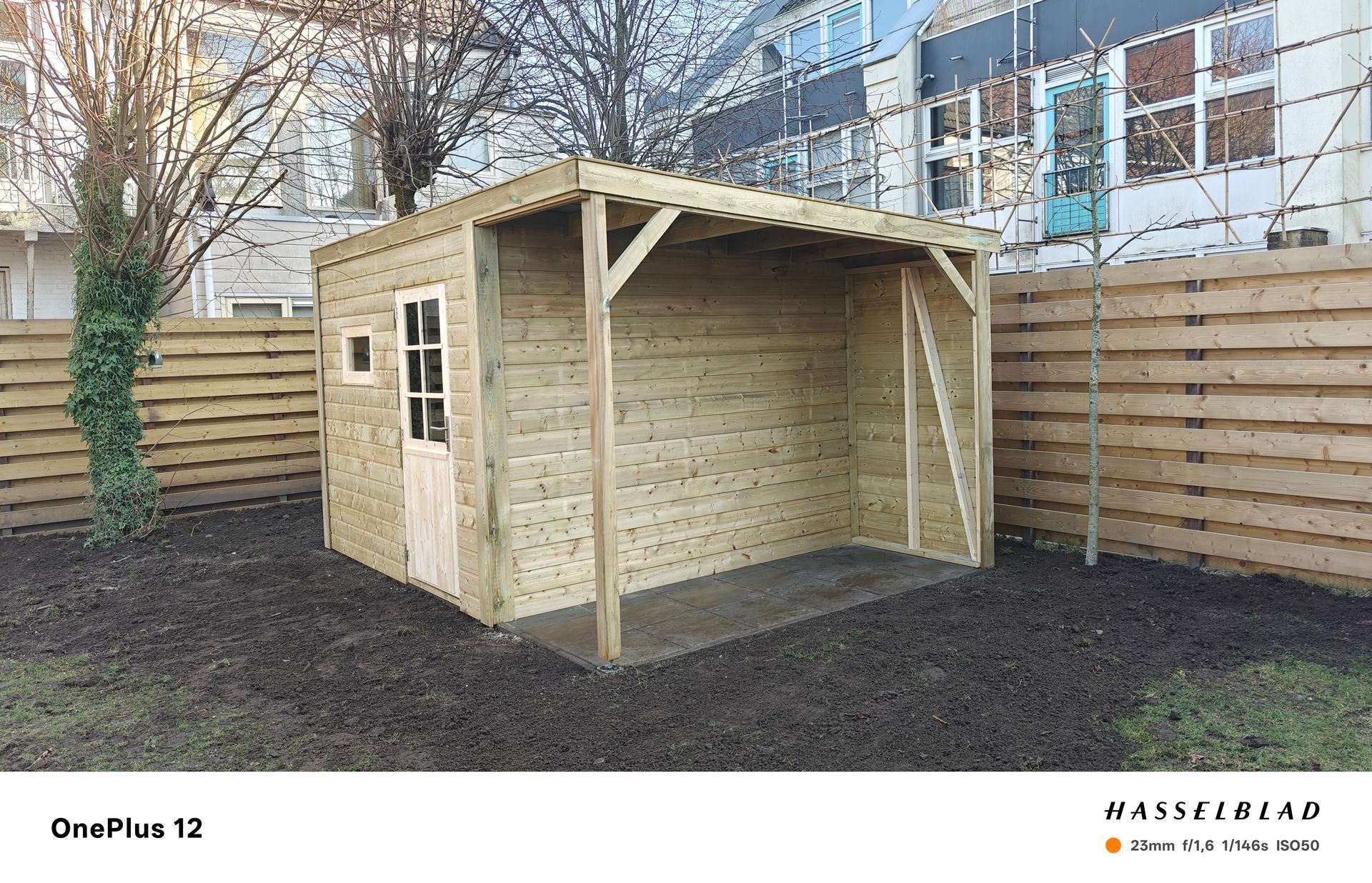 Houten schuur met een overdekte veranda in een tuin, naast een houten schutting.