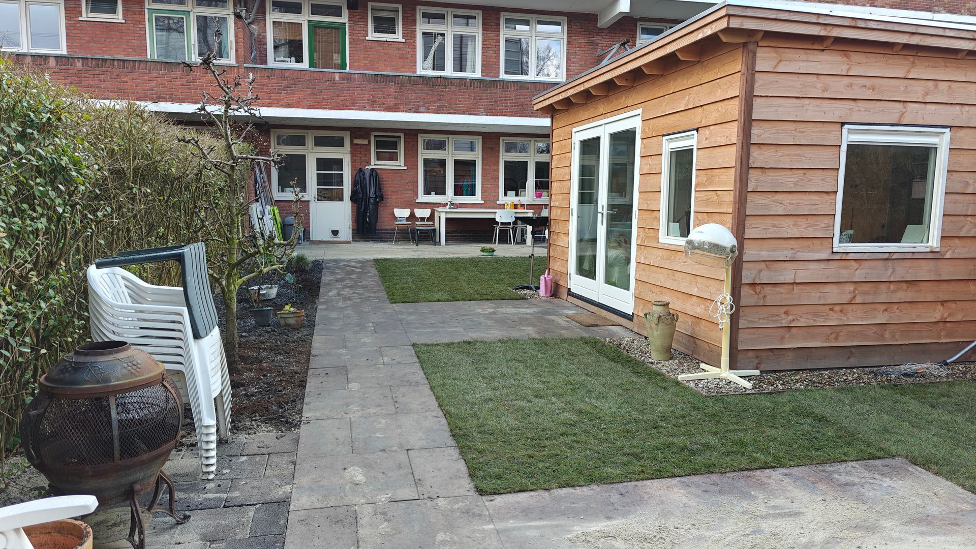 Achtertuin met een houten schuur, een bestraat pad, gras en een bakstenen gebouw op de achtergrond.