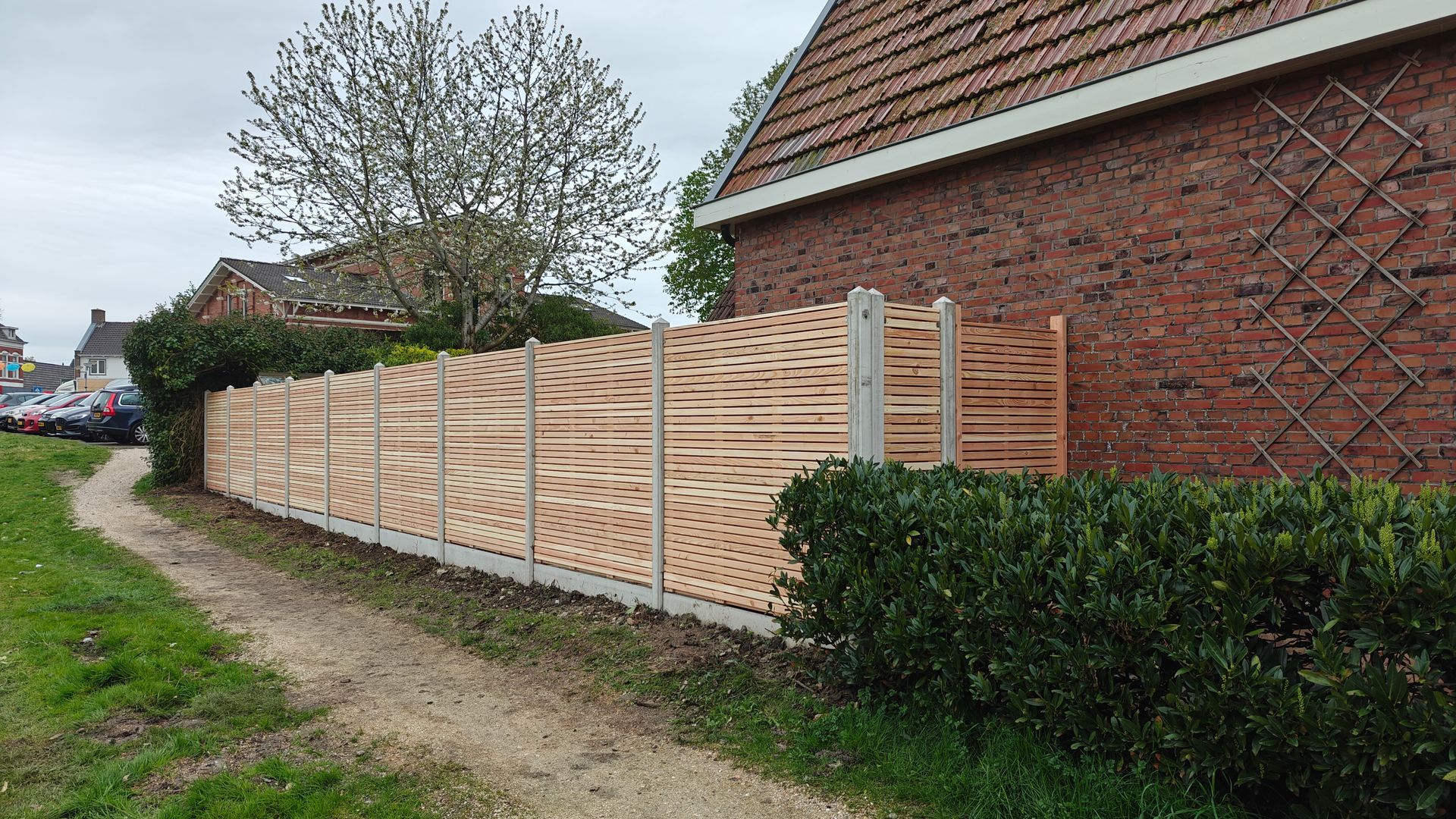 Houten schutting met patroon naast een bakstenen gebouw en een grindpad.