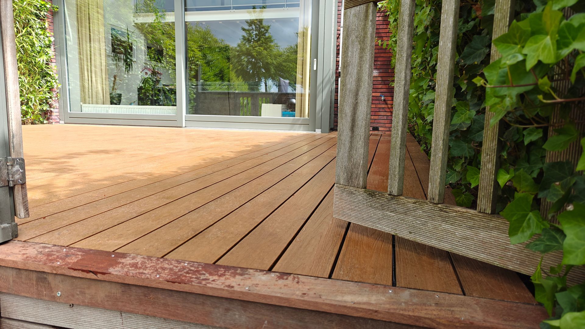 Een houten terras dat leidt naar glazen deuren, omgeven door groen en een verweerde balustrade.