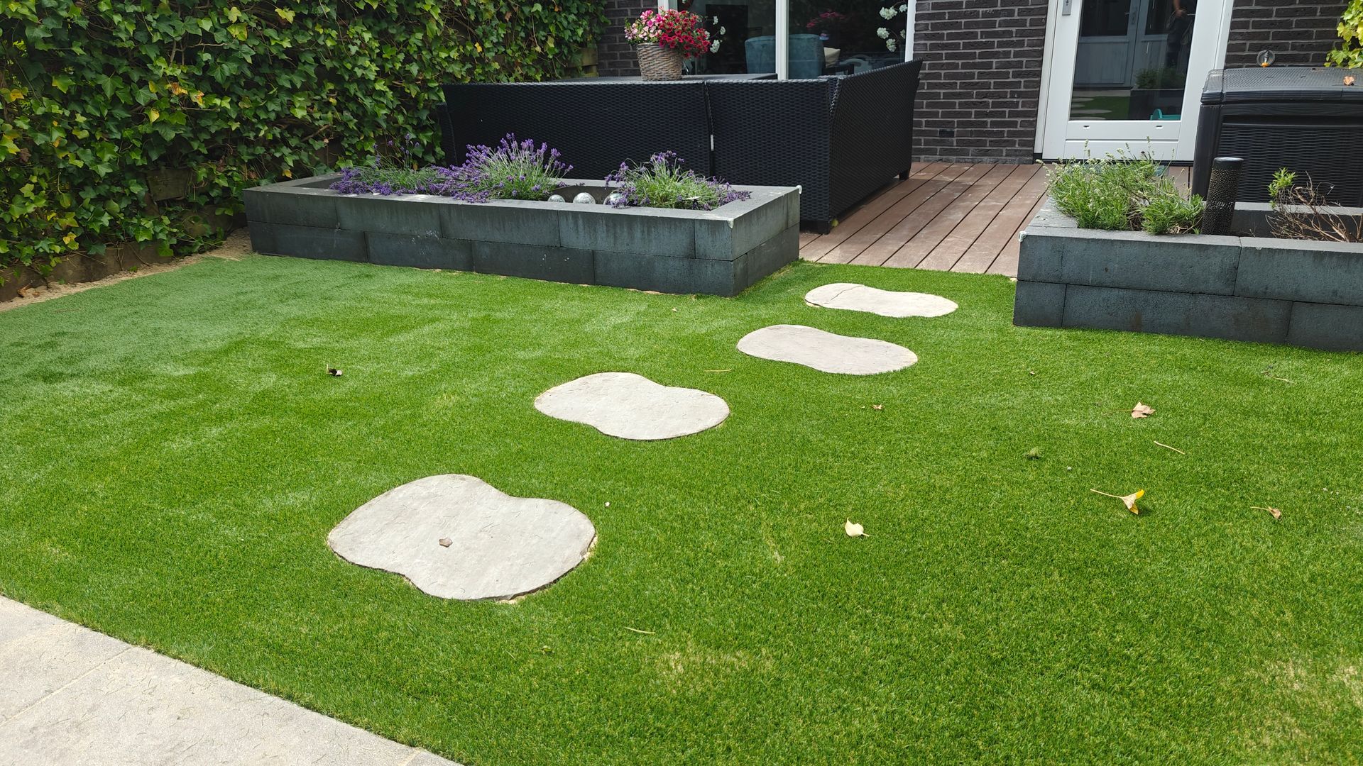 Groen gazon met stapstenen die leiden naar een terras met bloembakken en een zitgedeelte.