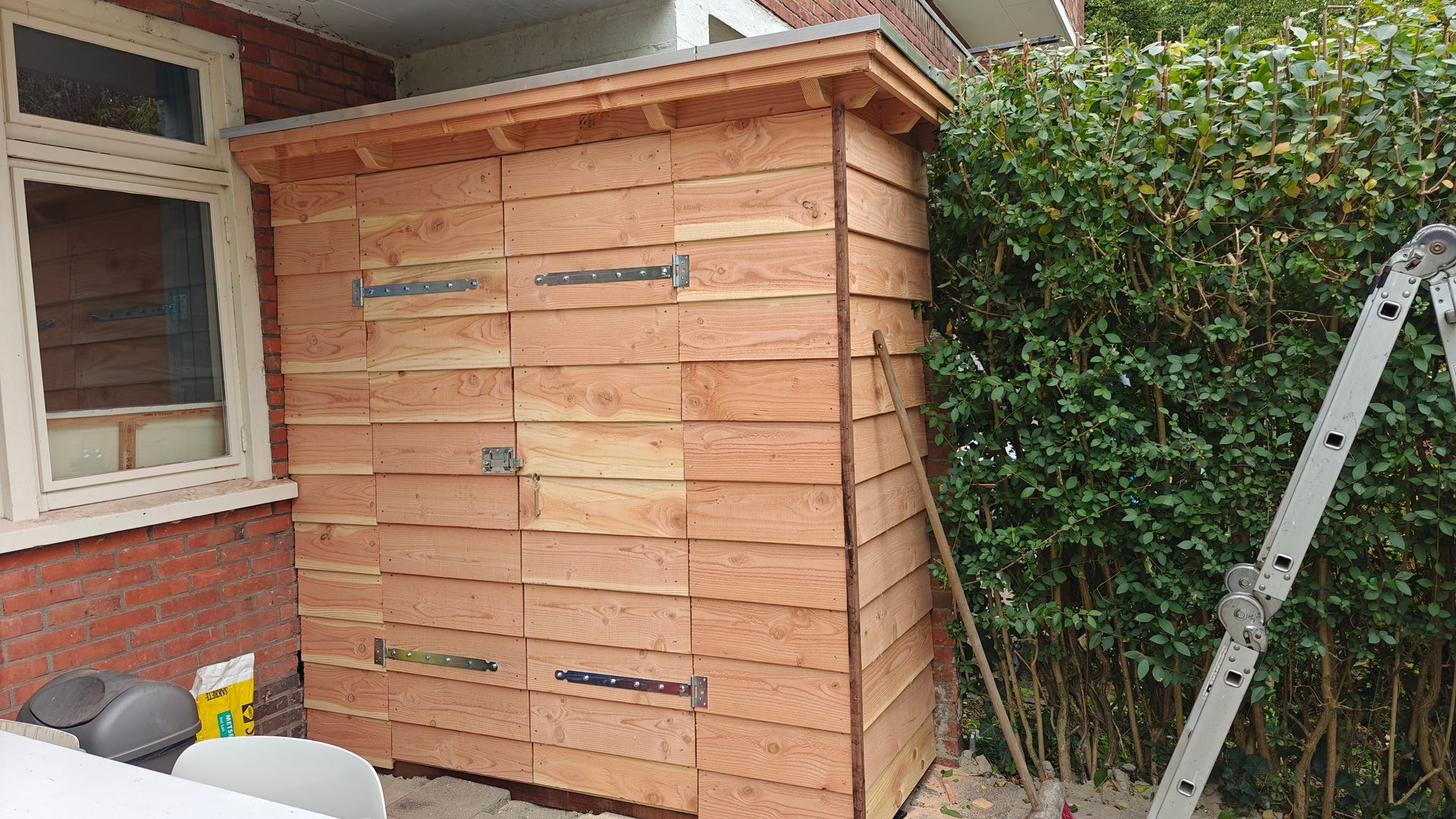 Een houten schuur staat tegen een gebouw, naast een struik. Een ladder leunt tegen de schuur.