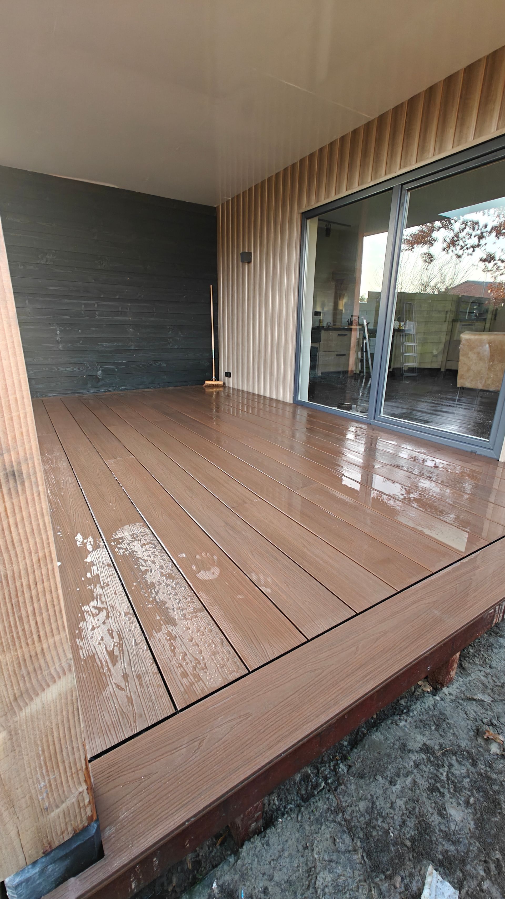 Houten terras met een nat oppervlak, omgeven door houten lambrisering en een glazen schuifdeur.