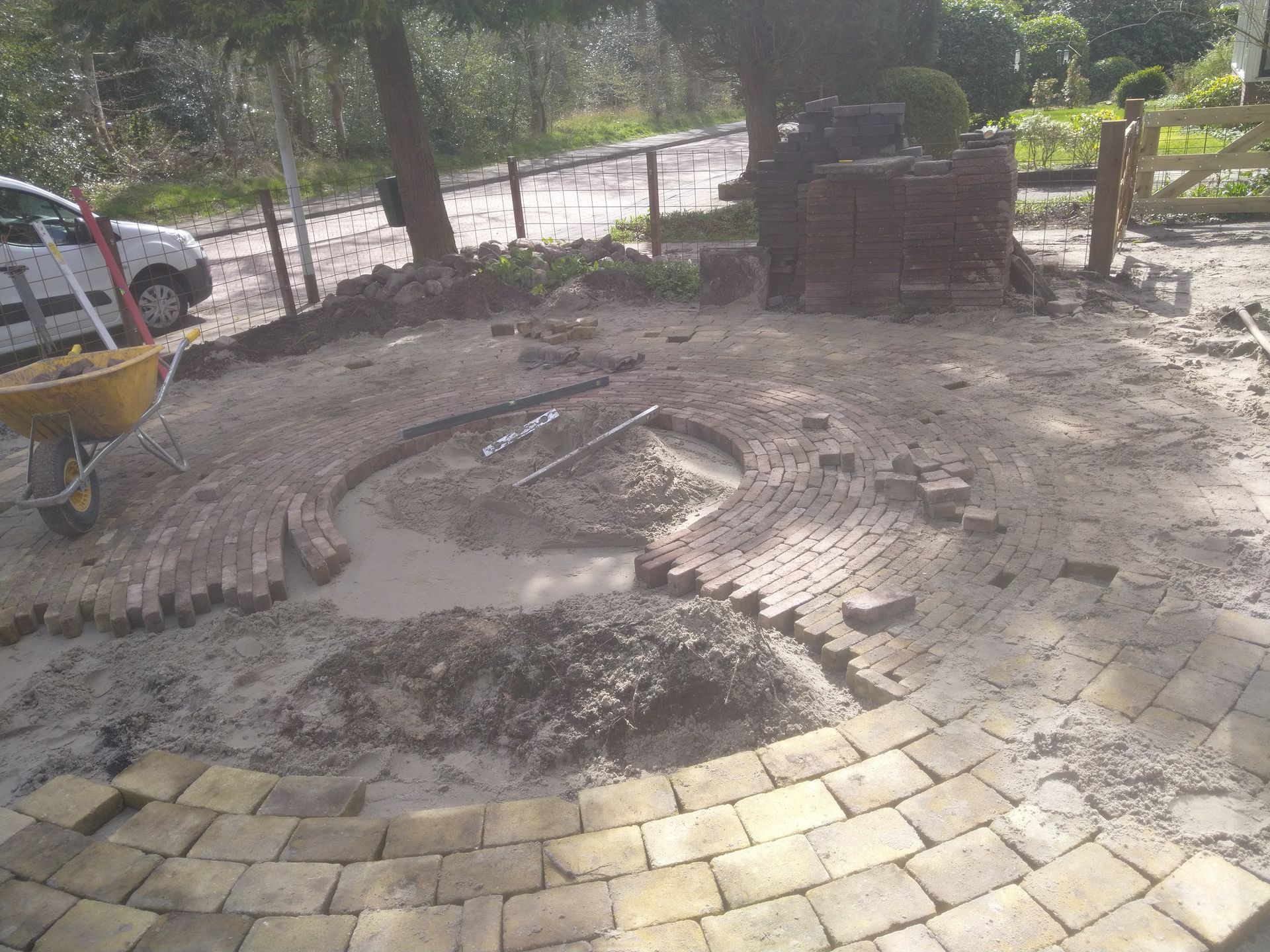 Metselwerk in uitvoering, cirkelvormig ontwerp, mortel en bakstenen op zandgrond met een kruiwagen en auto op de achtergrond.