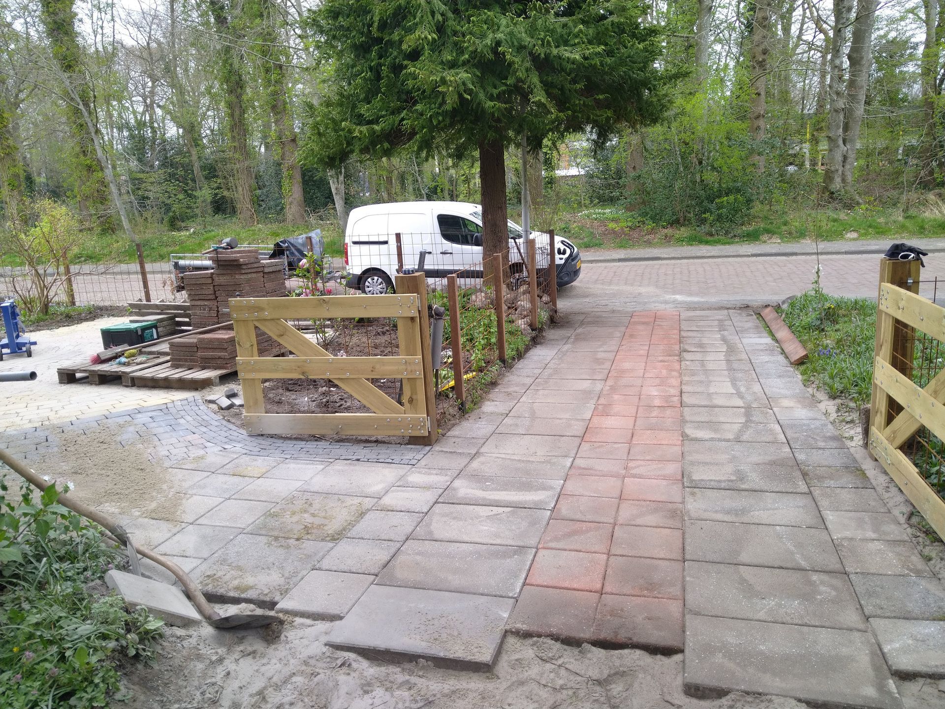 Pad in aanbouw met bestrating, zand, houten poort en een geparkeerde bestelwagen.