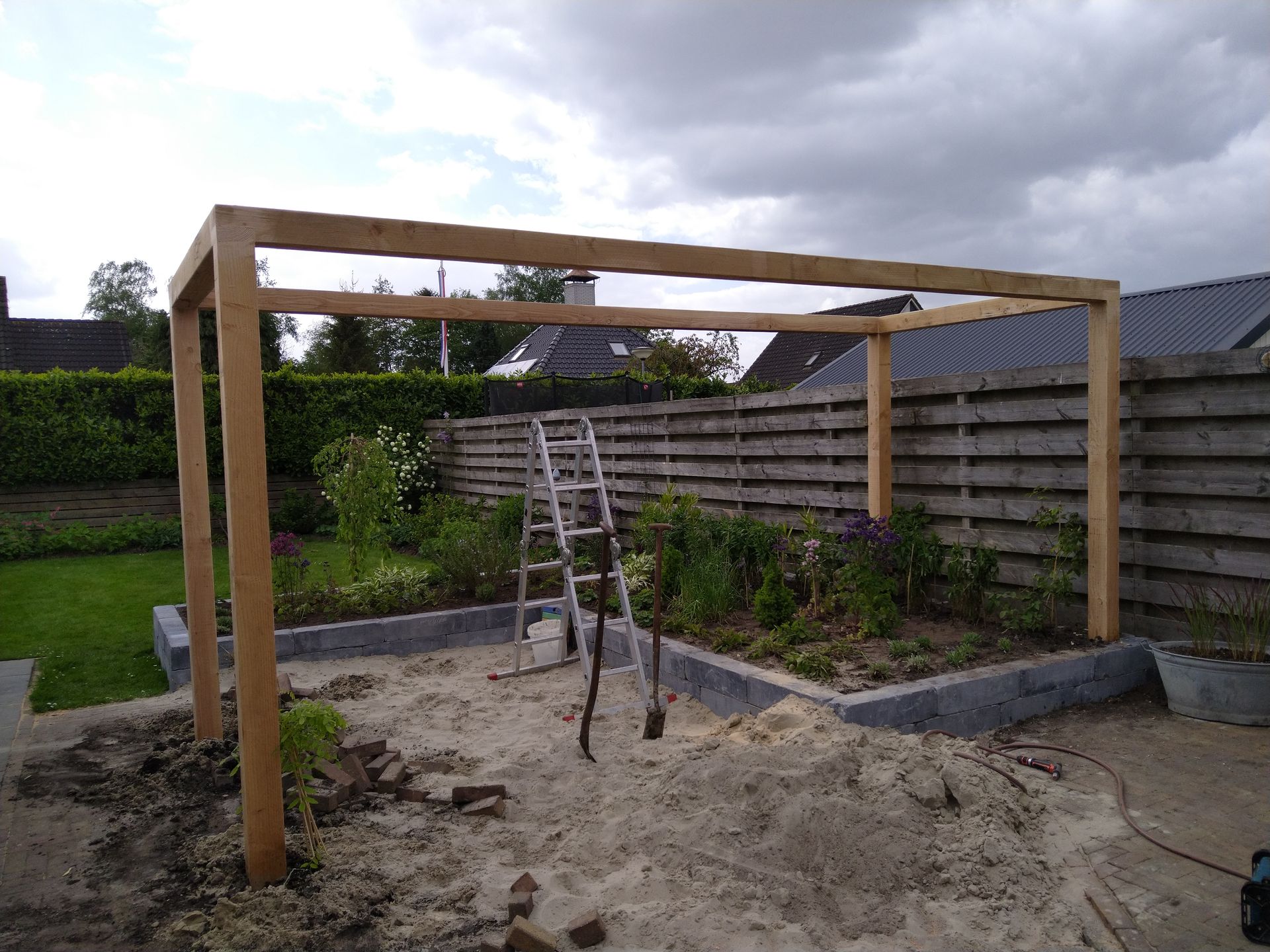 Houten pergola in aanbouw in een tuin, ladder aanwezig, bewolkte lucht.