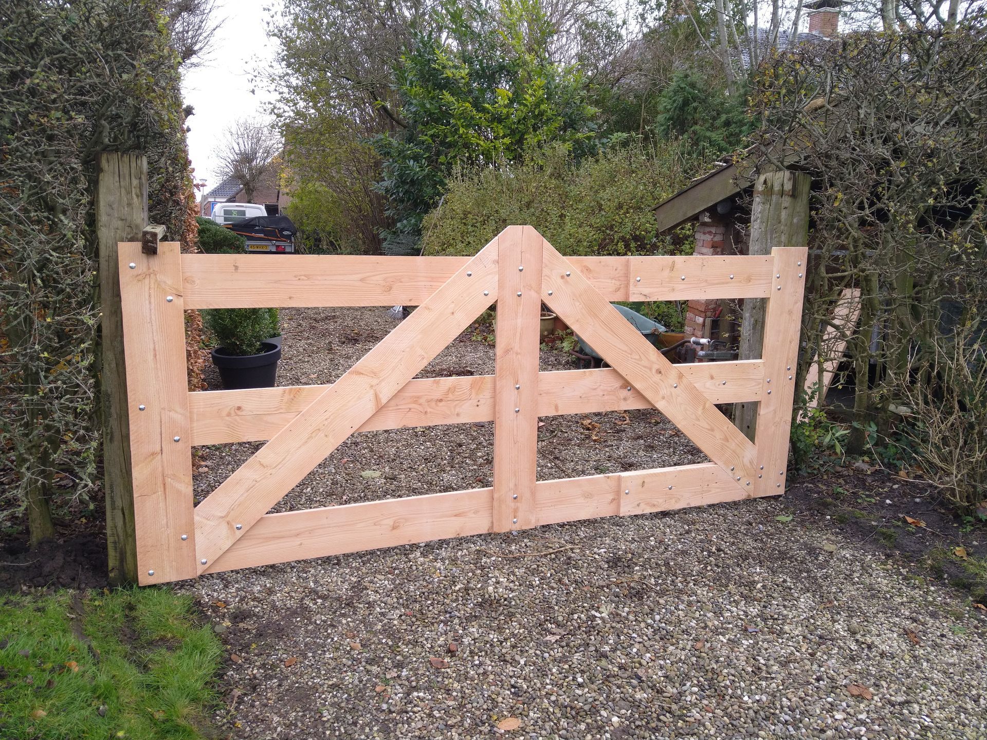 Houten poort met diagonale verstevigingen, die uitkomt op een grindpad, geflankeerd door hagen en palen.