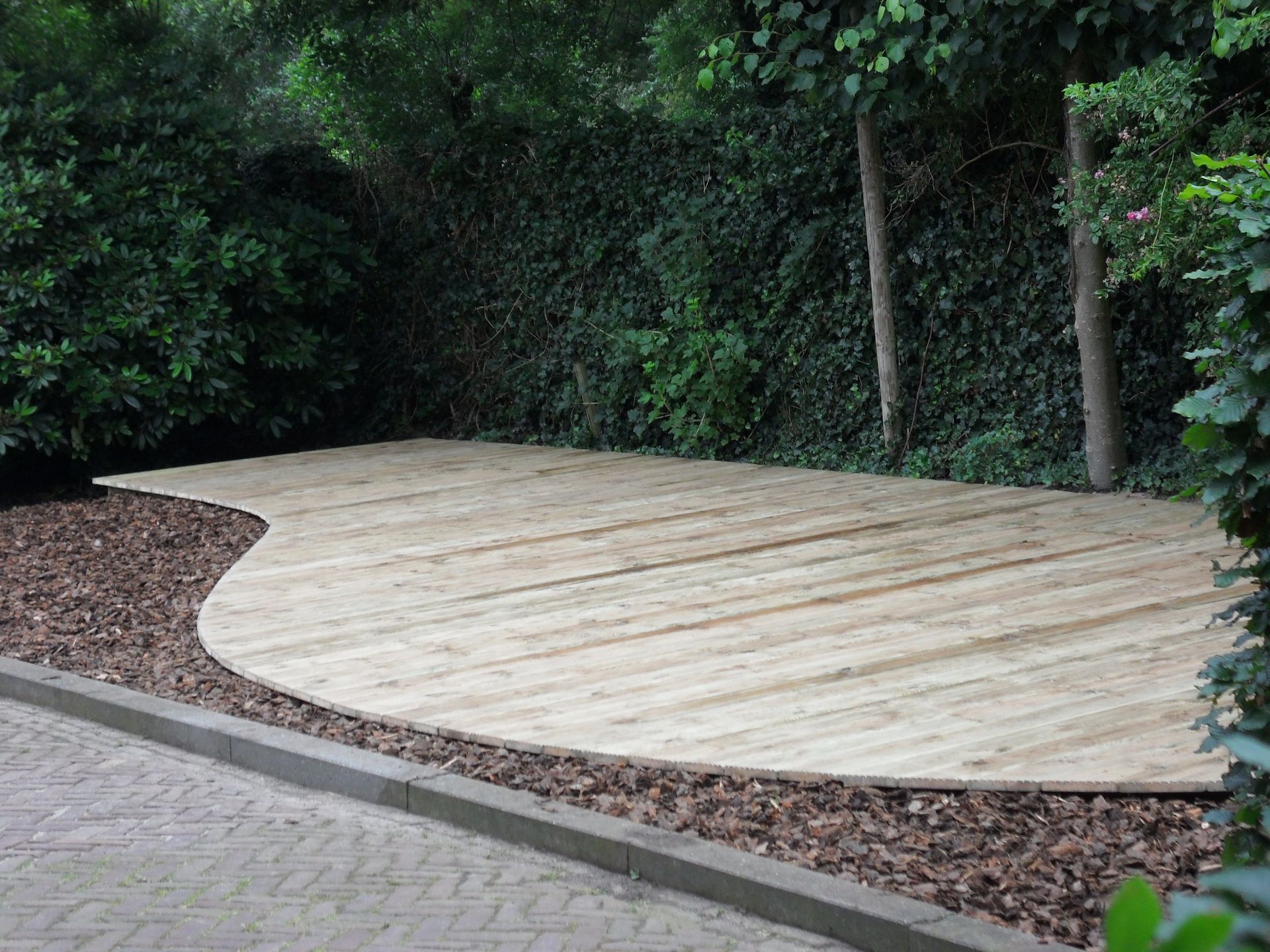 Houten terras in een tuin, omgeven door bomen en houtsnippers, naast een bestraat gedeelte.