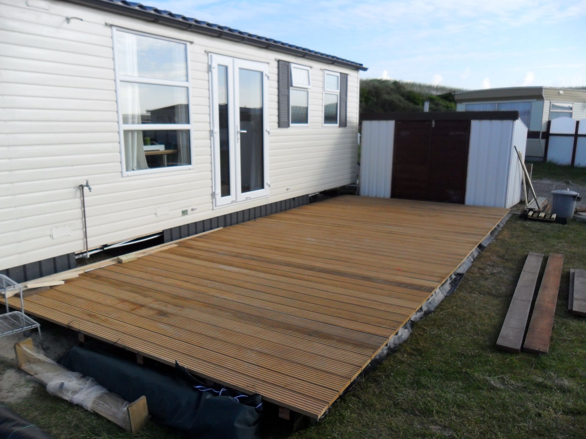 Houten terrasuitbouw naast een beige stacaravan. Het terras is bruin en staat op gras.