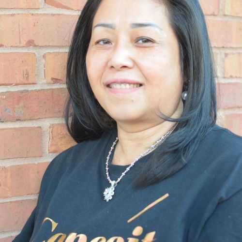 Woman with black hair and a black shirt smiles, leaning against a red brick wall. She wears a necklace.