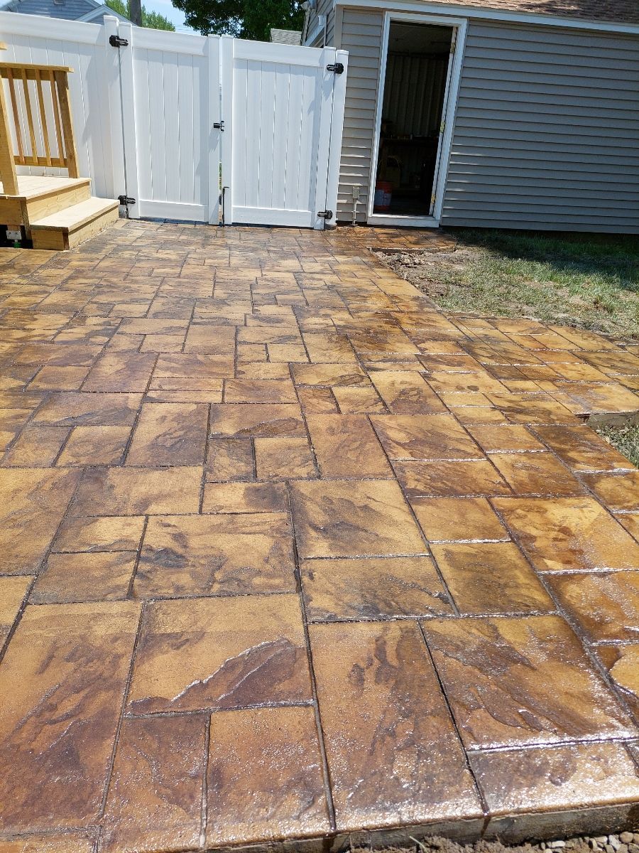 Stamped concrete patio with a brown and tan stone pattern leads to a white gate and a small shed.