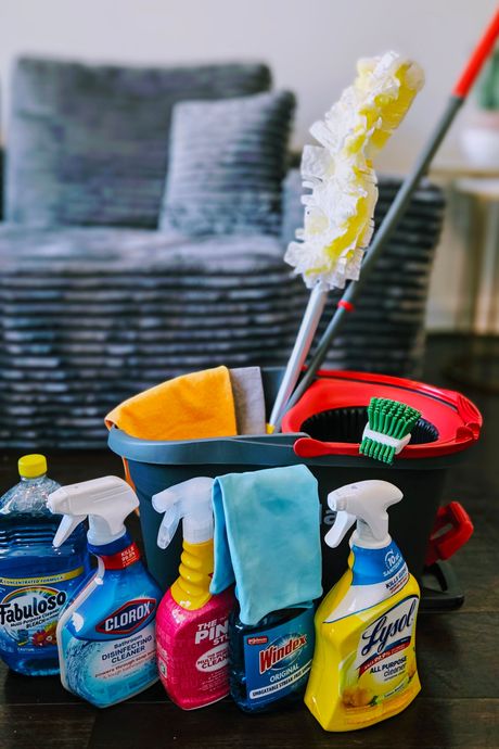 Cleaning supplies, including spray bottles, cloths, bucket, mop, and duster, on a dark wood floor near a sofa.