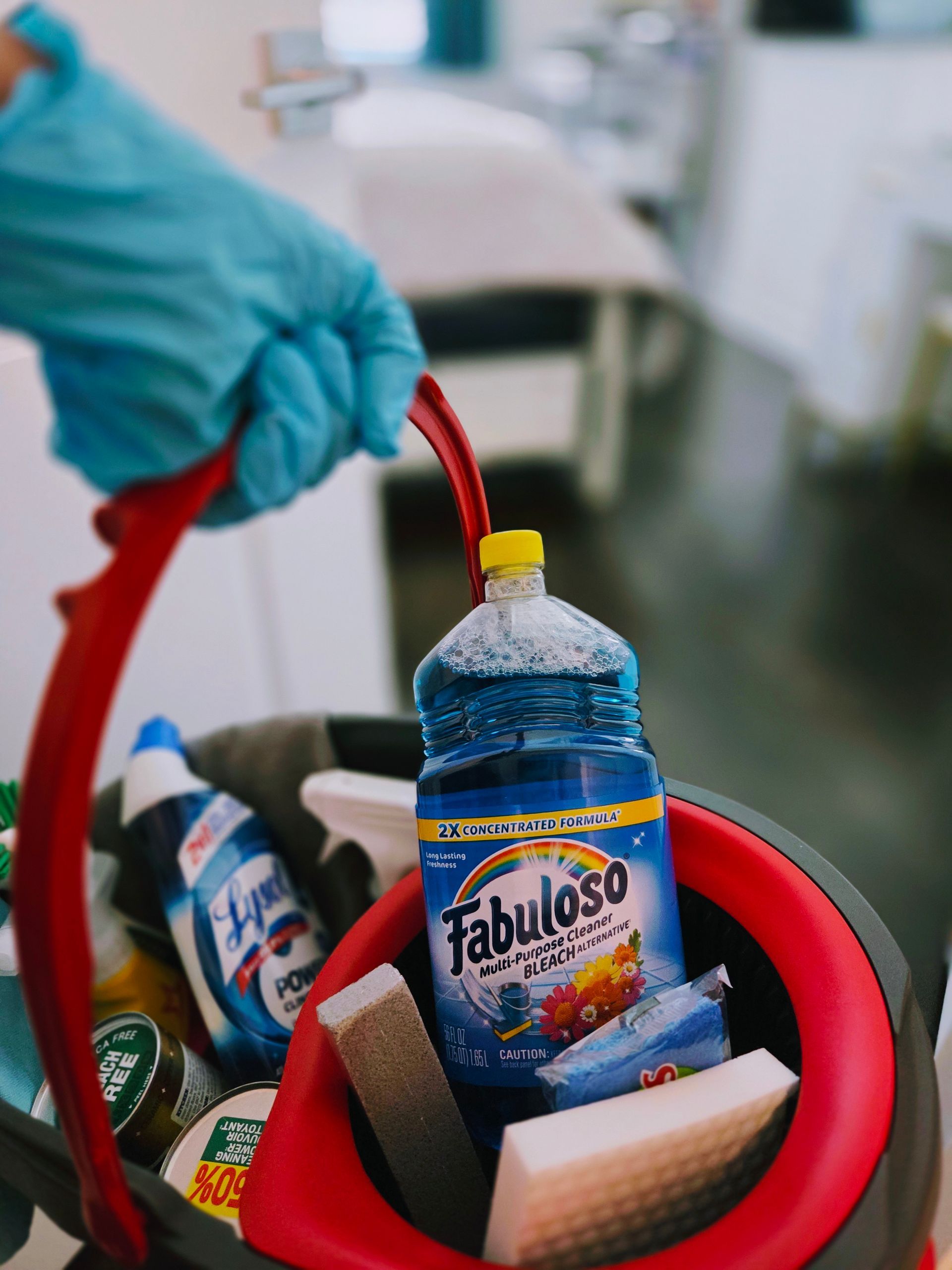 A hand in blue glove holds a red cleaning caddy filled with cleaning supplies.