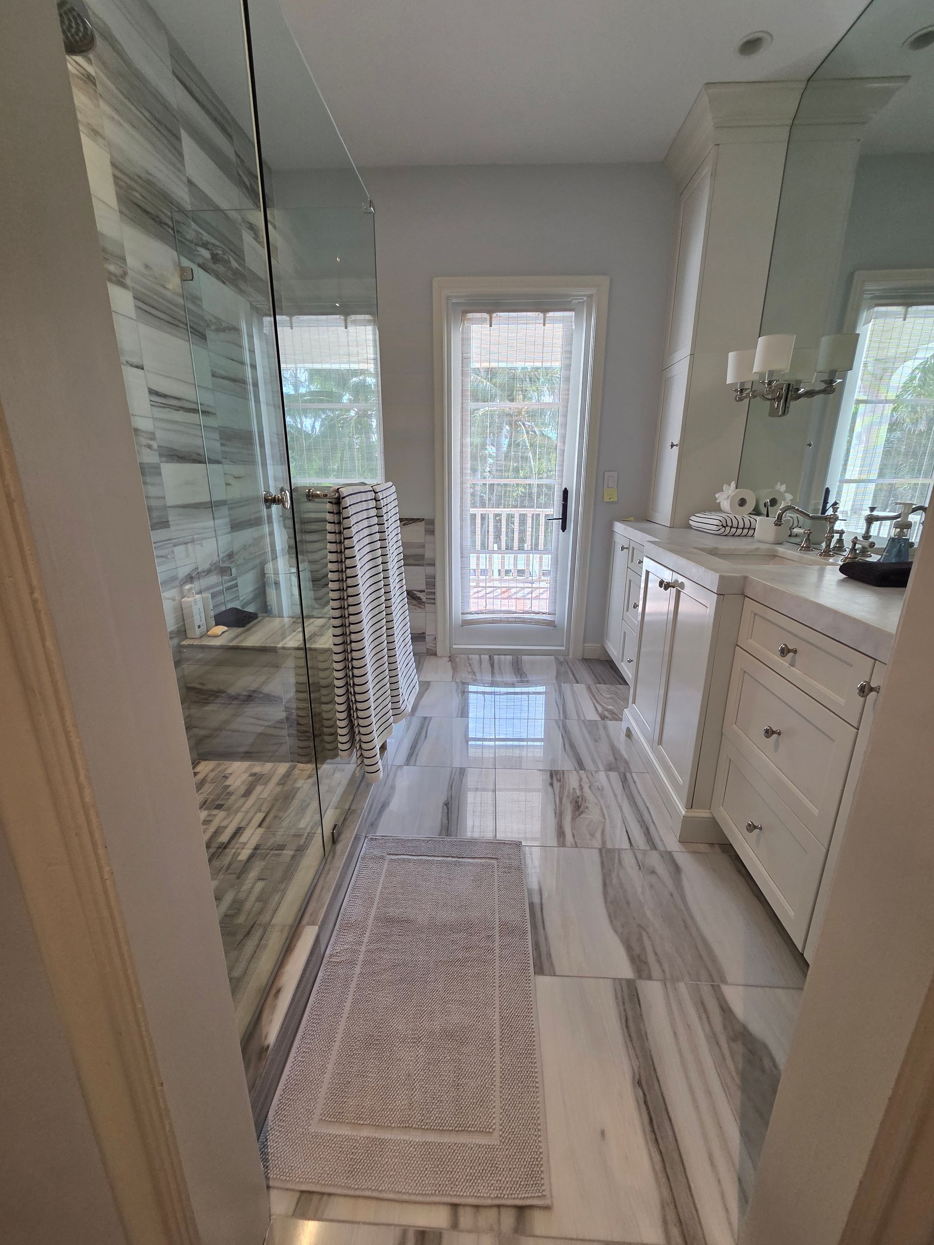 Bathroom with marble floor and walls, white vanity, glass shower, and door to outside.