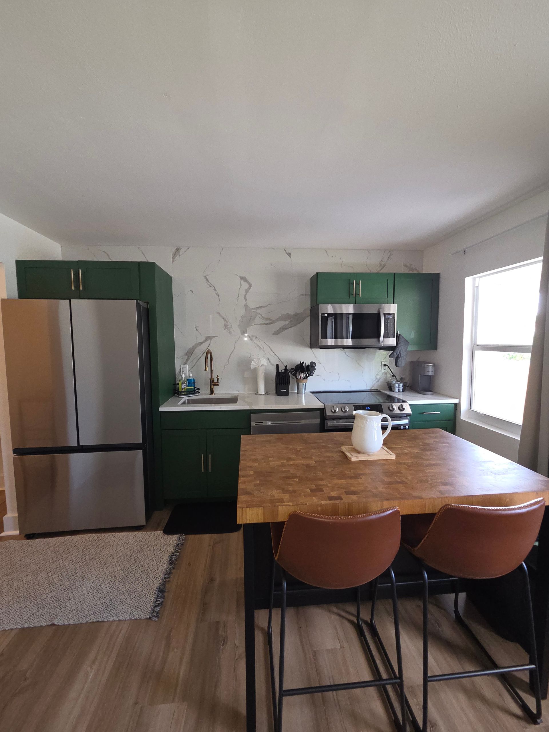 Green kitchen with stainless steel appliances, butcher block island, and two brown chairs.